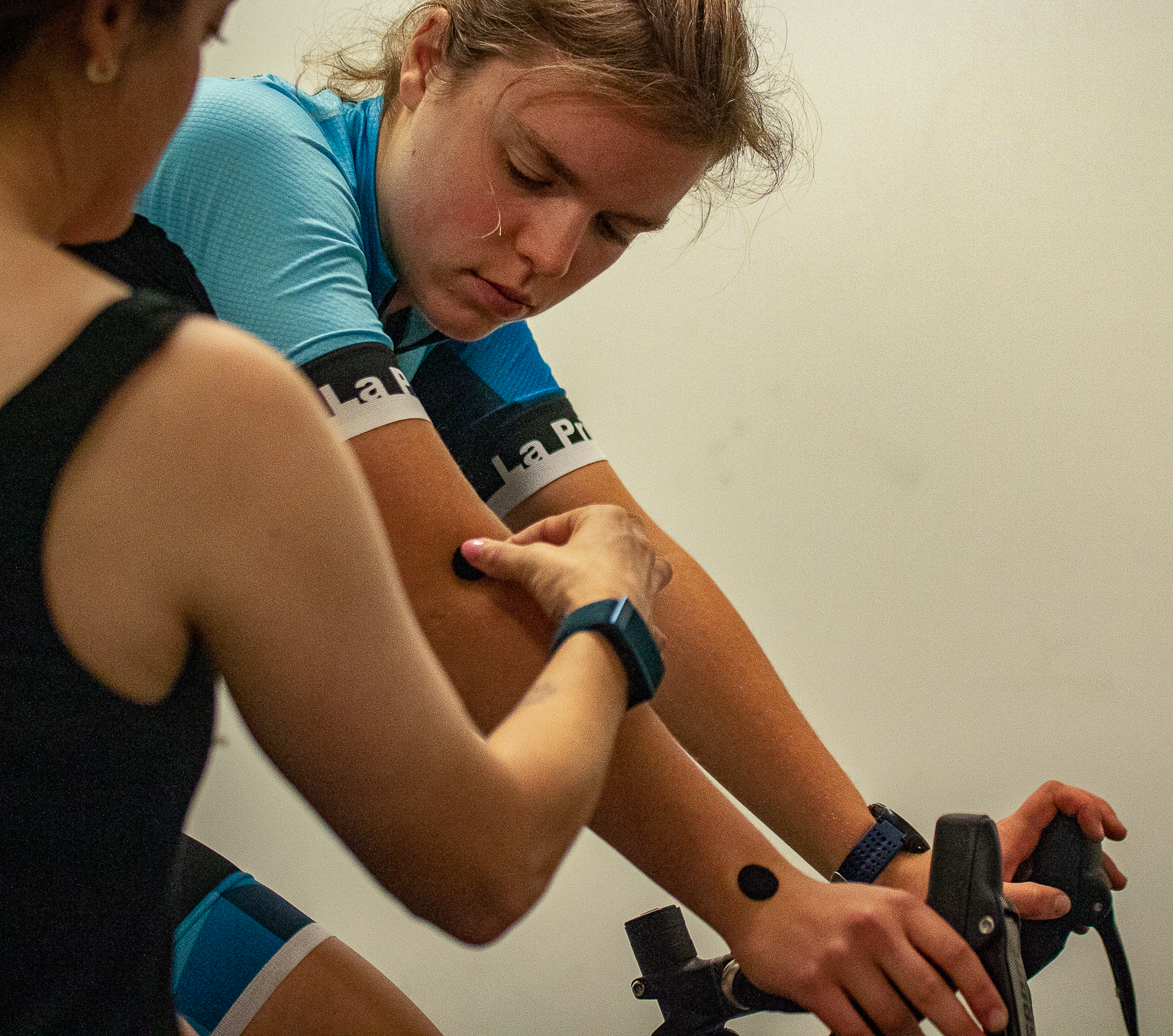 Woman in blue cycling gear having black circular patches applied to her arms while holding bicycle handlebars.