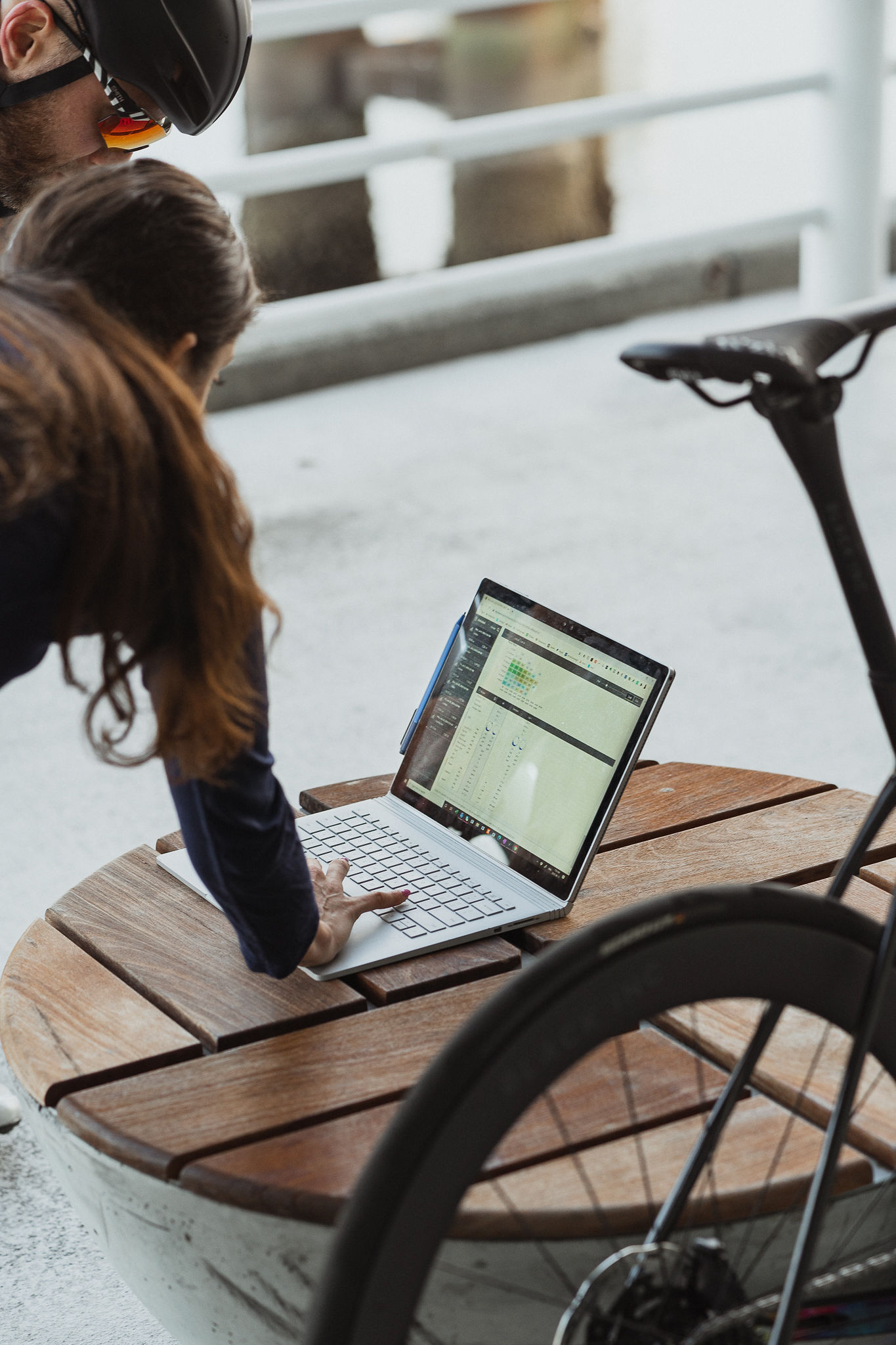 Two people looking at a laptop on a wooden outdoor table next to a bicycle wheel.