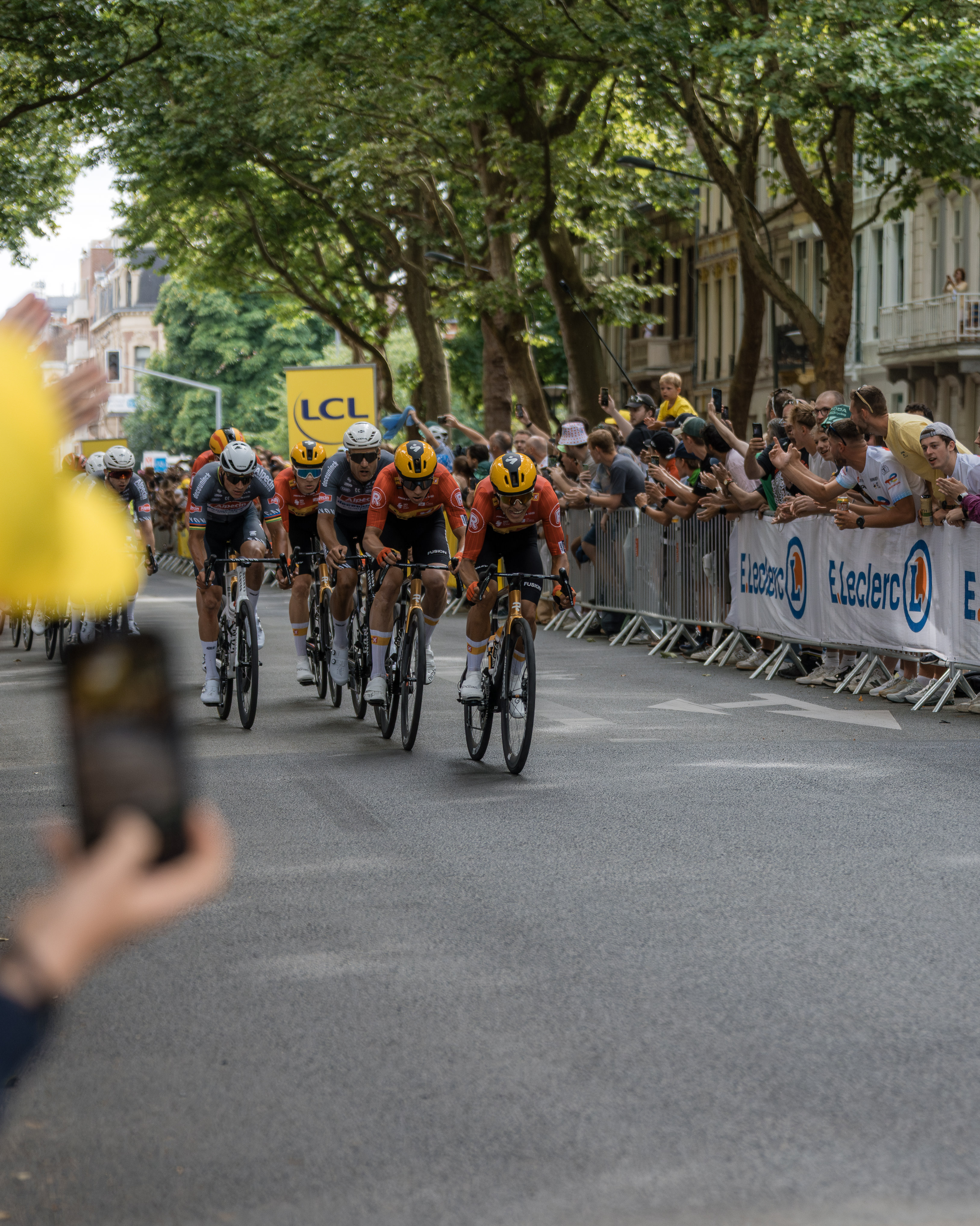 Cyclists in orange and black jerseys racing on a tree-lined street with cheering crowds behind barriers.