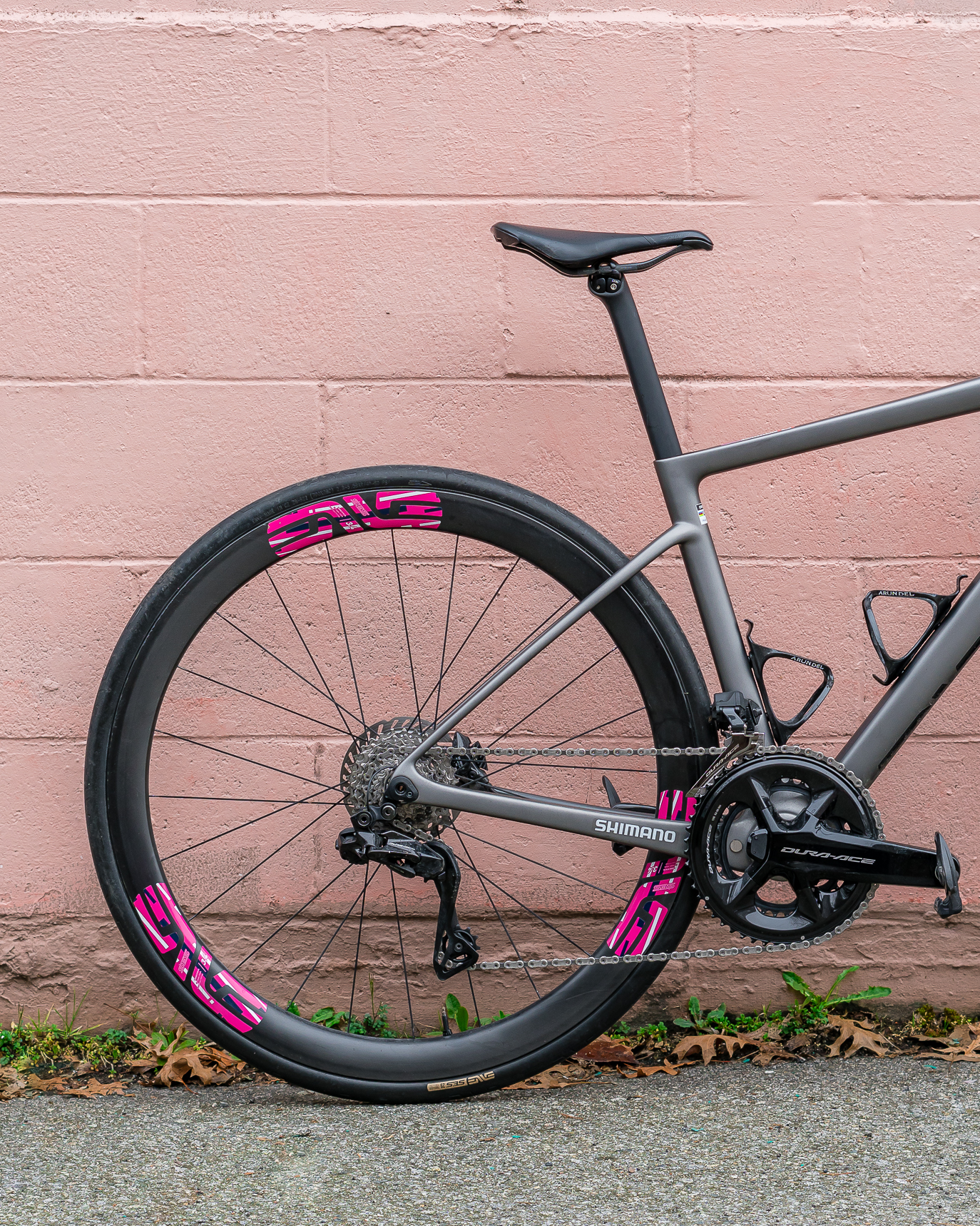 Rear half of a matte gray road bike with deep carbon rims, pink ENVE decals, Shimano drivetrain, and black saddle against a pink brick wall.