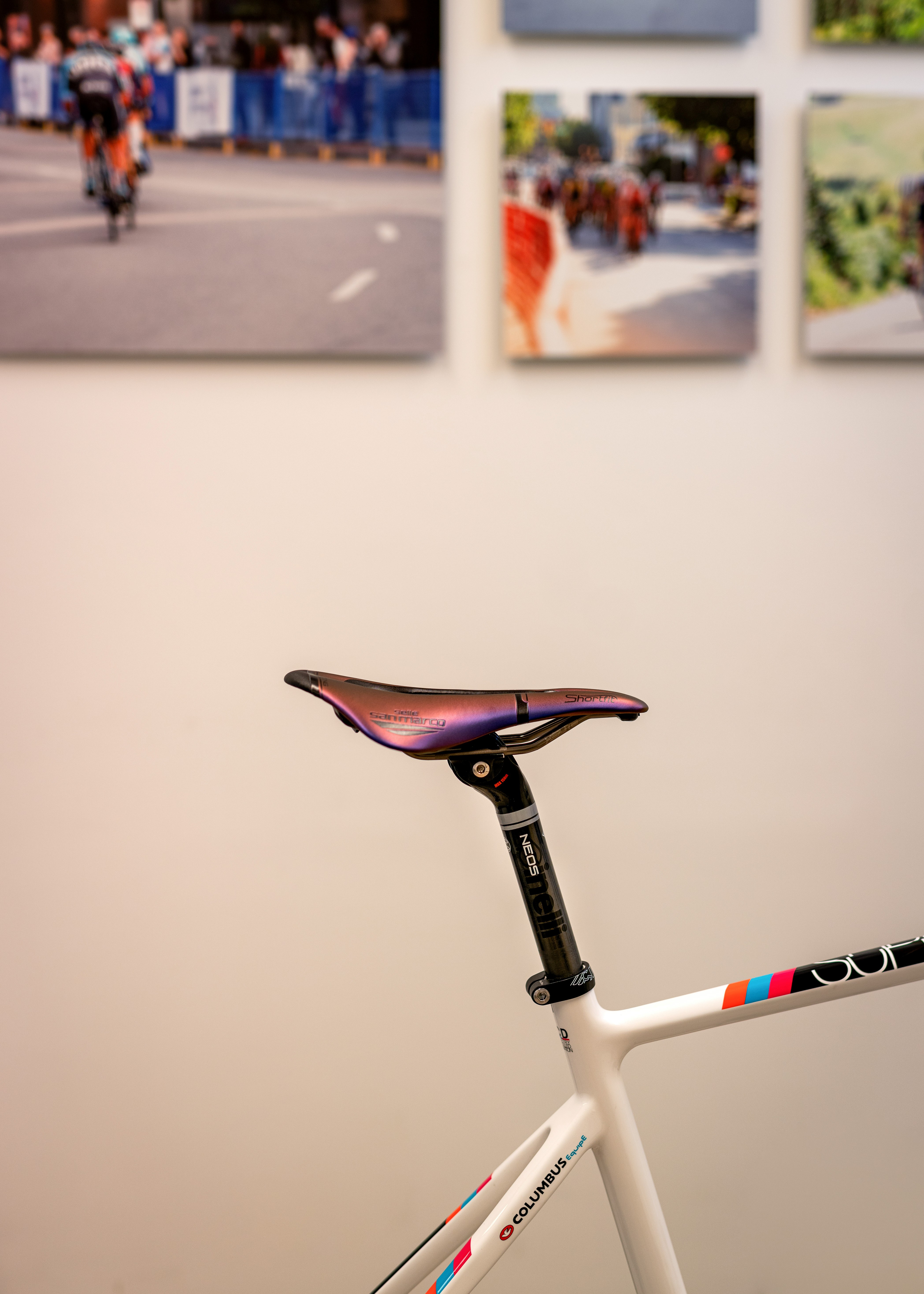 Close-up of a white bicycle frame with colorful stripes and a purple Selle San Marco saddle against a blurred background of cycling race photos on the wall.