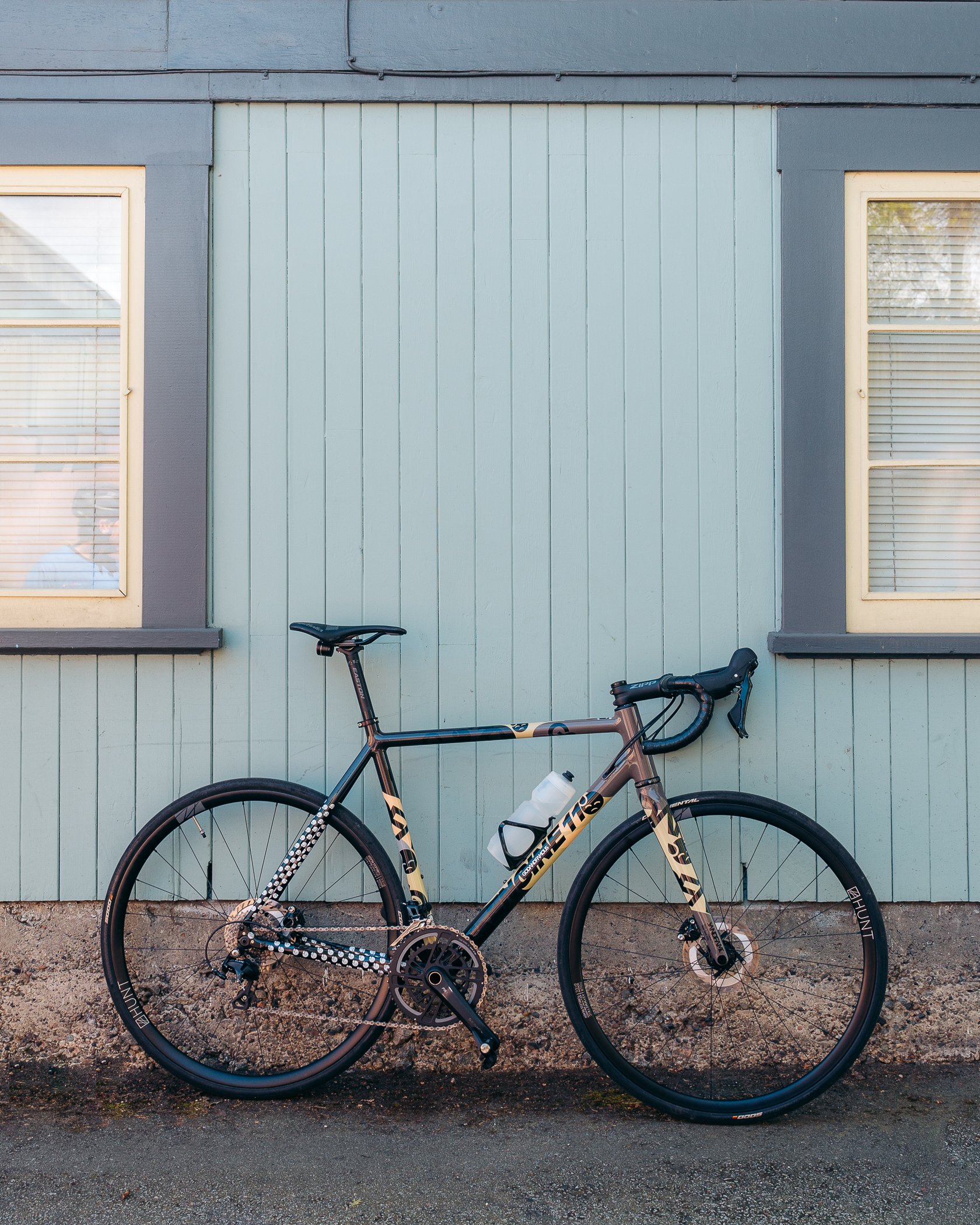 Road bike with black and beige frame leaning against a light blue wooden wall with two windows.