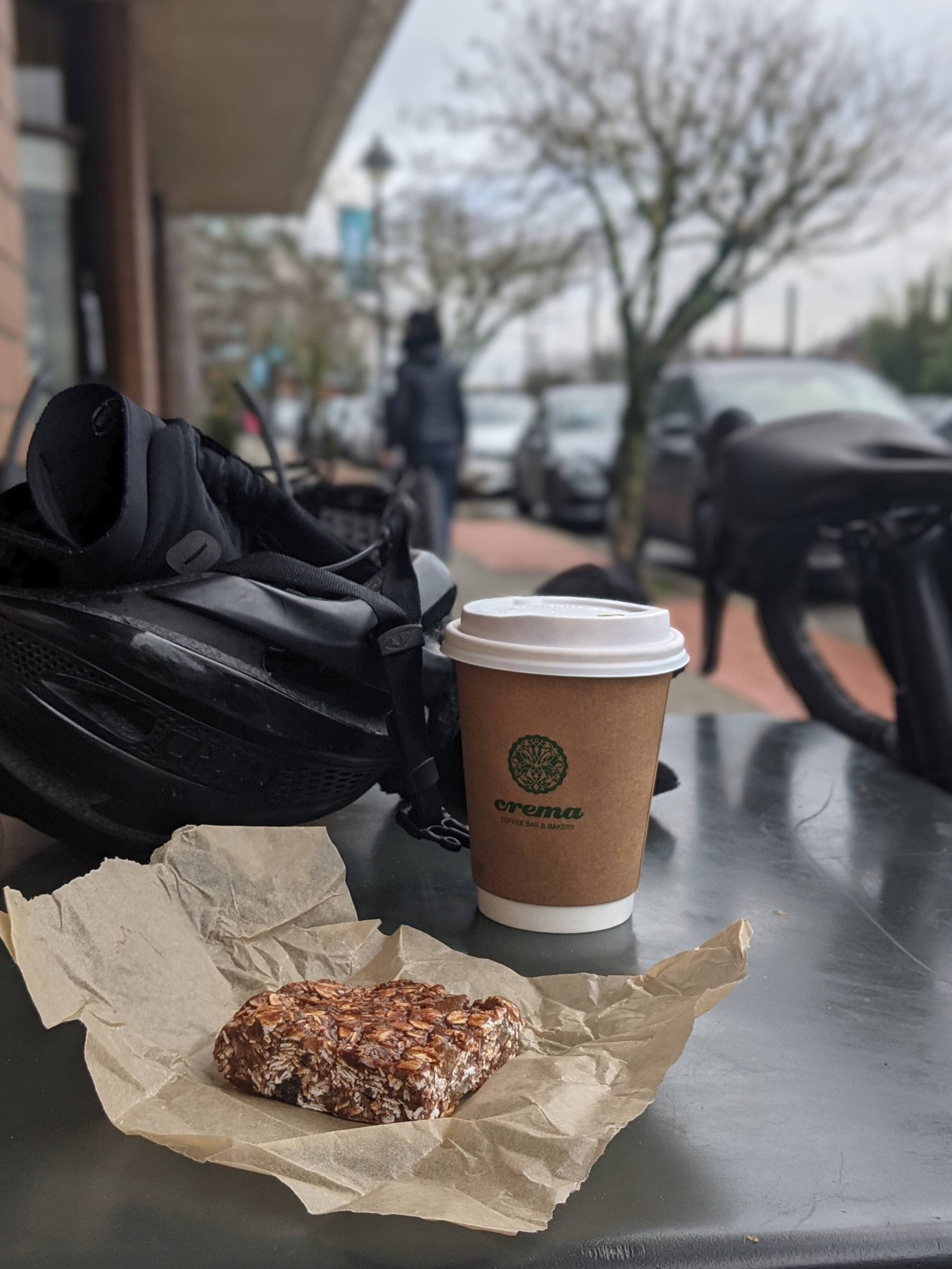Granola bar on parchment paper next to a coffee cup with 'Crema Coffee Bar & Bakery' logo on a black table, with a black bicycle helmet and handlebar in the background.
