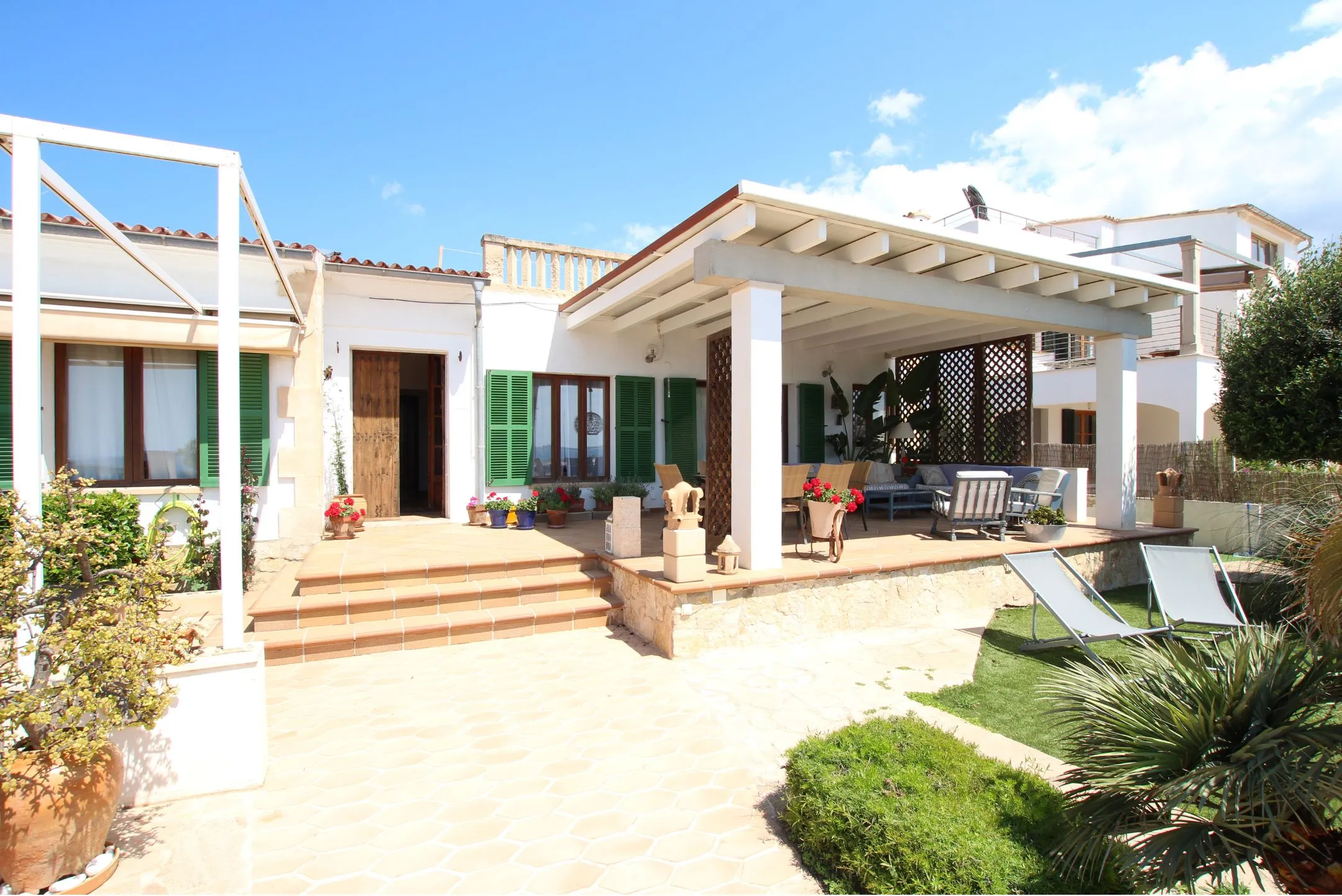 Sunlit outdoor patio area with terracotta tiled steps, white pergola, green shuttered windows, potted plants, and two lounge chairs on grass.