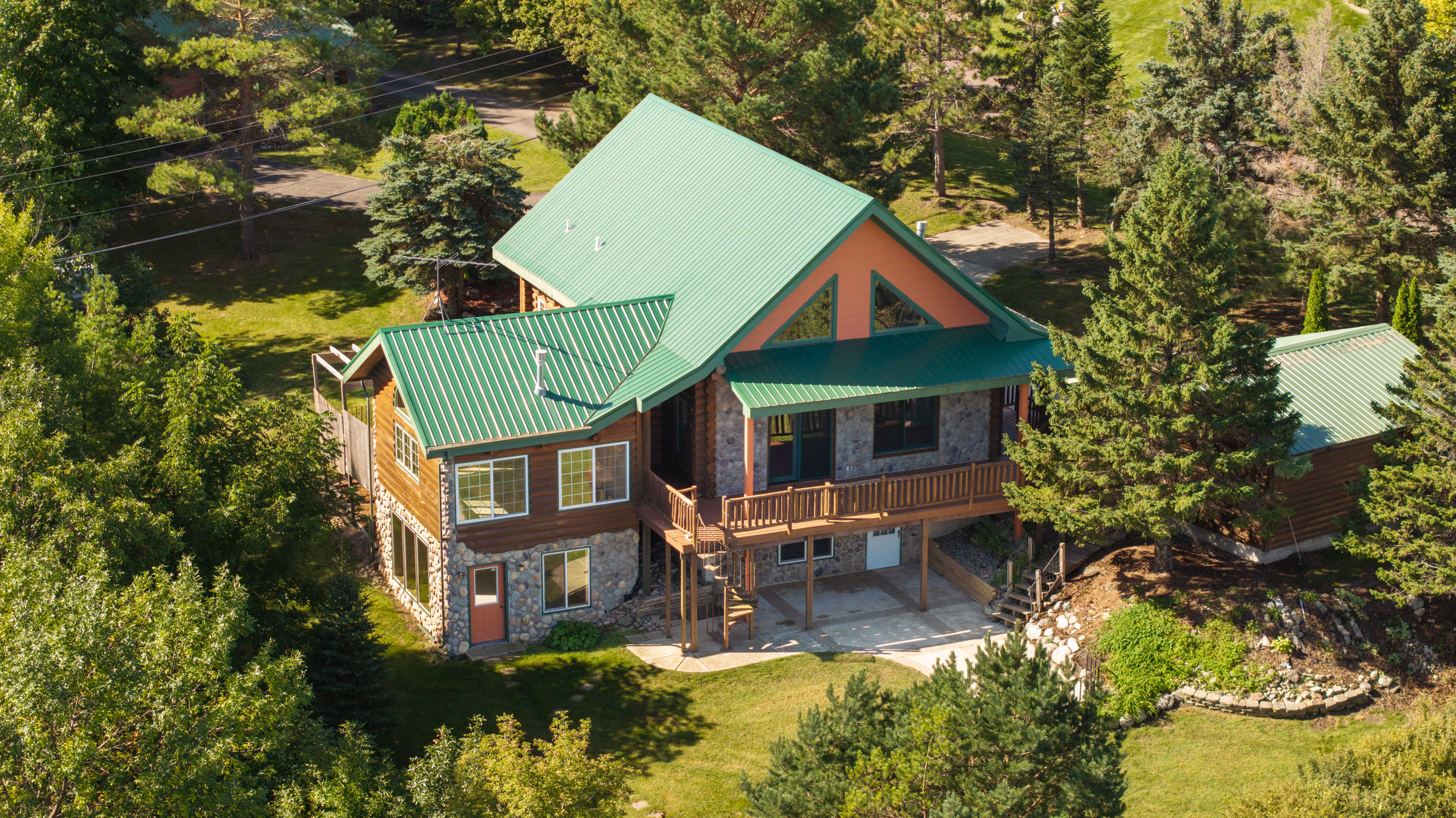 A large rustic house with a green metal roof surrounded by dense green trees and lawn, featuring stone and wood exterior, a covered porch with wooden railings, and a spiral staircase leading down to a concrete patio.