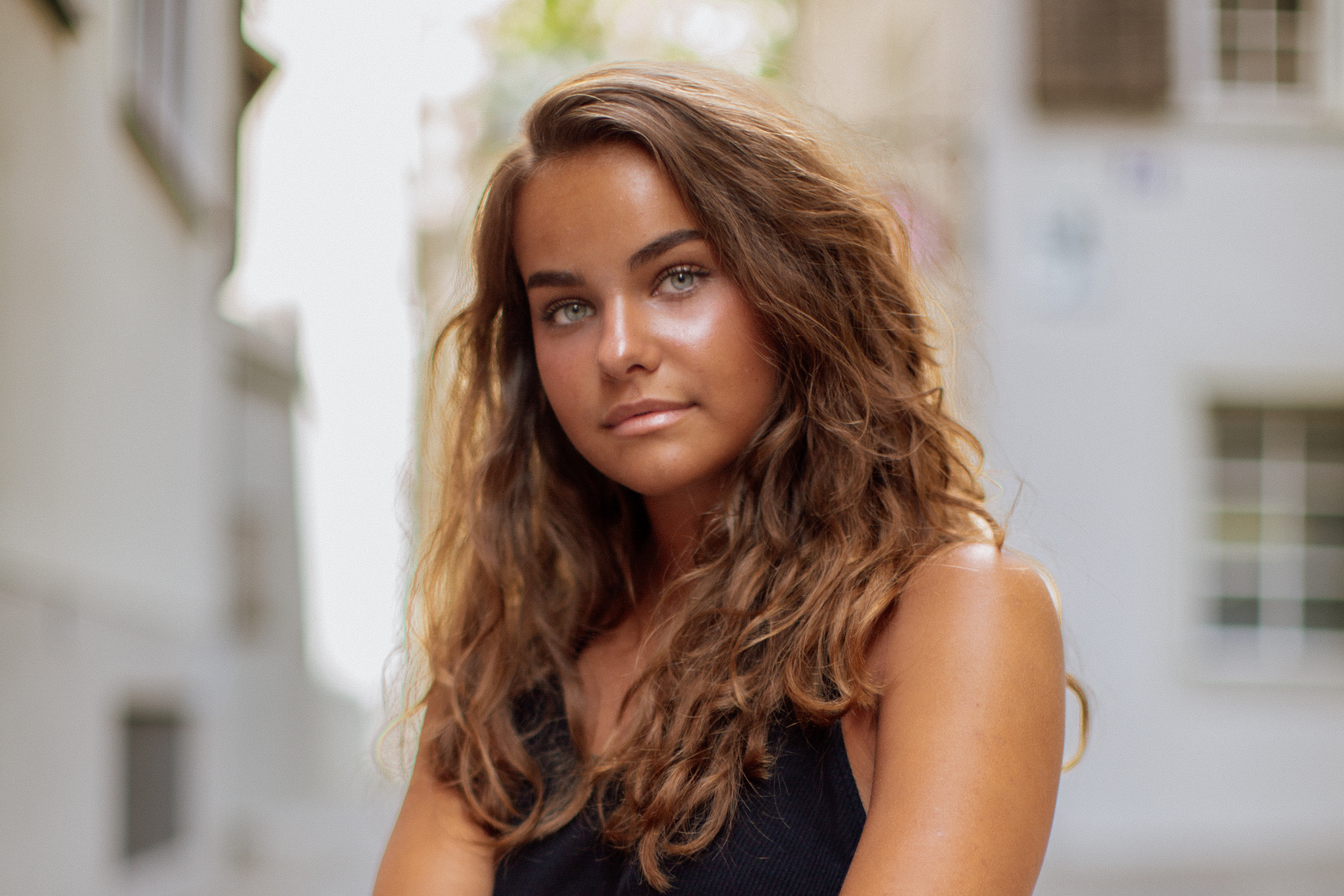 Young woman with long wavy brown hair and green eyes wearing a black sleeveless top, looking slightly to the right against a blurred outdoor background.