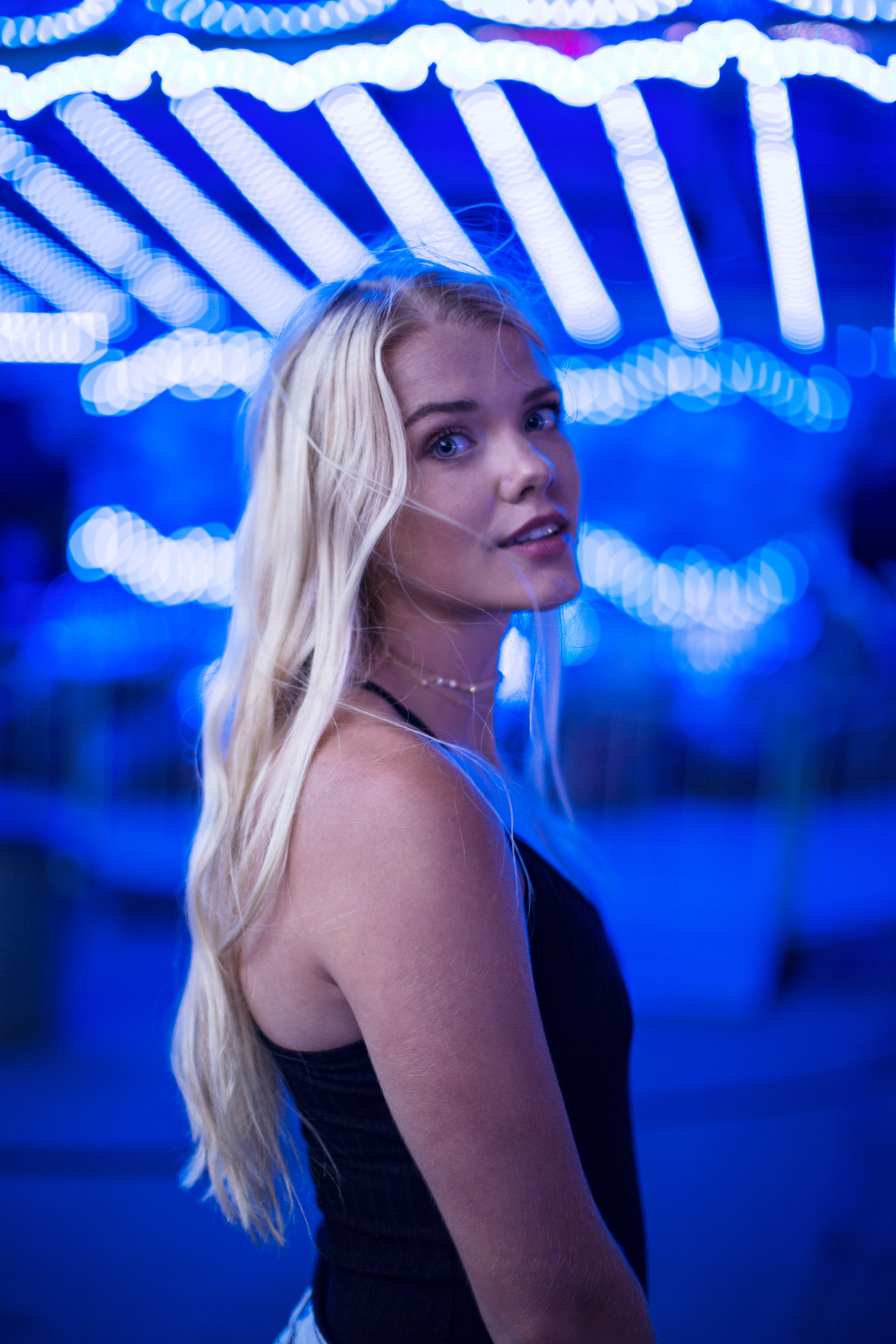 Young woman with long blonde hair wearing a black sleeveless top stands turned slightly with her head looking back over her shoulder, illuminated by blue string lights above and behind her at night.