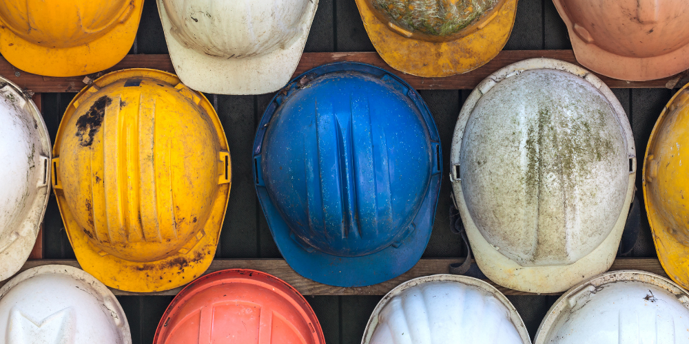 Various worn construction helmets in yellow, white, blue, orange, and beige hanging on a wall.
