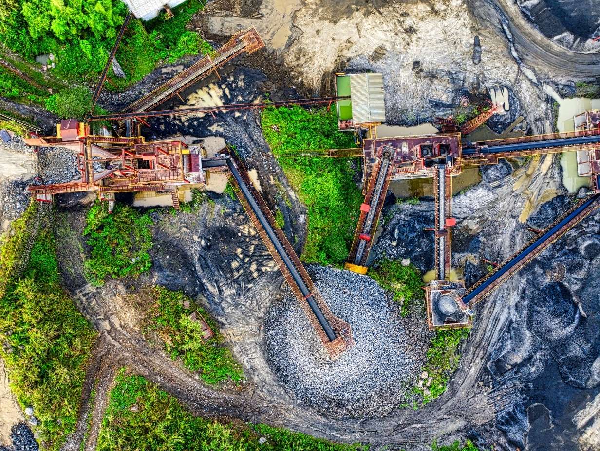 Aerial view of an industrial quarry site with conveyor belts transporting gravel and rocks surrounded by greenery and muddy terrain.