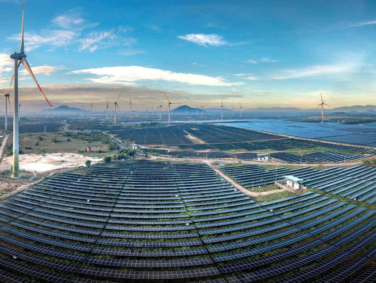 Wide solar farm with rows of solar panels and multiple wind turbines under a blue sky with scattered clouds.