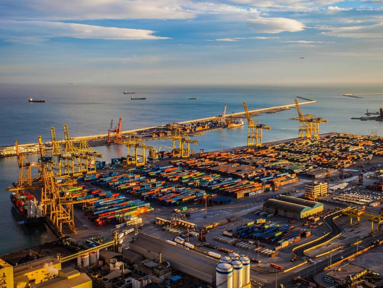 Aerial view of a busy shipping port with numerous colorful cargo containers, large yellow cranes, ships docked, and the ocean with several ships in the distance under a partly cloudy sky.