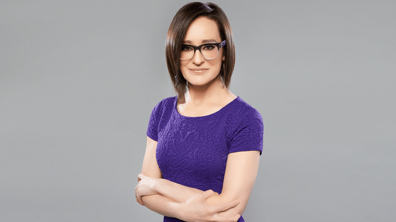 Kennedy wearing a purple patterned dress, standing with arms crossed against a gray background.