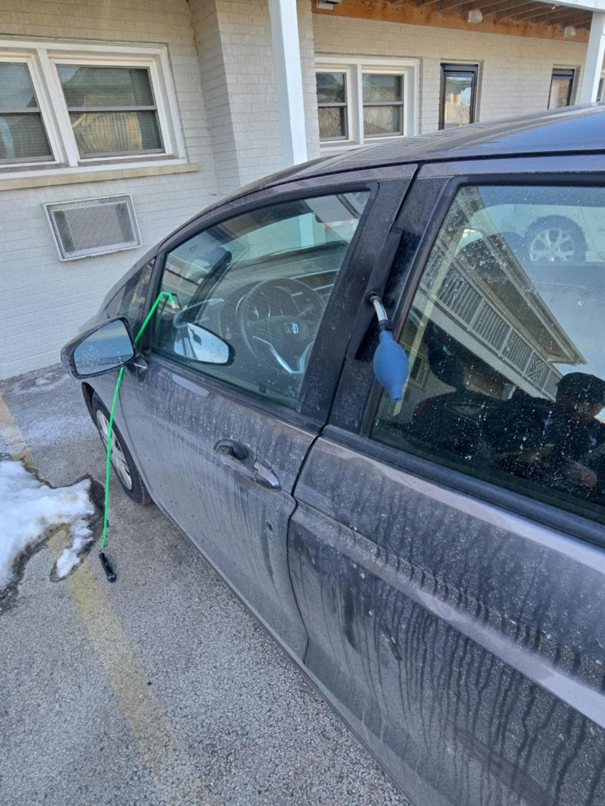 Dirty gray car parked outside with a green cable extending from the inside to outside, connected to side mirror and inflated blue bulb attached to car door frame.