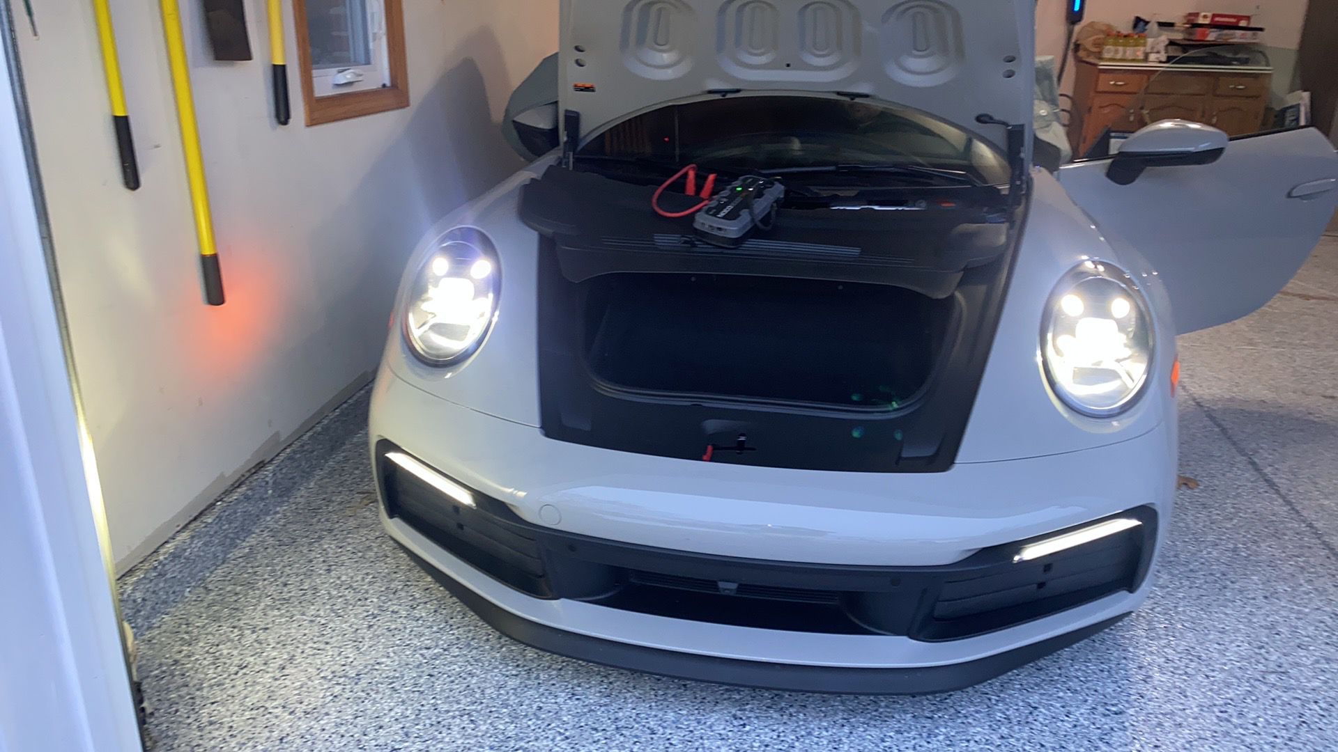 Front view of a white sports car with headlights on, front trunk open showing a battery charger inside, and right door open in a garage.