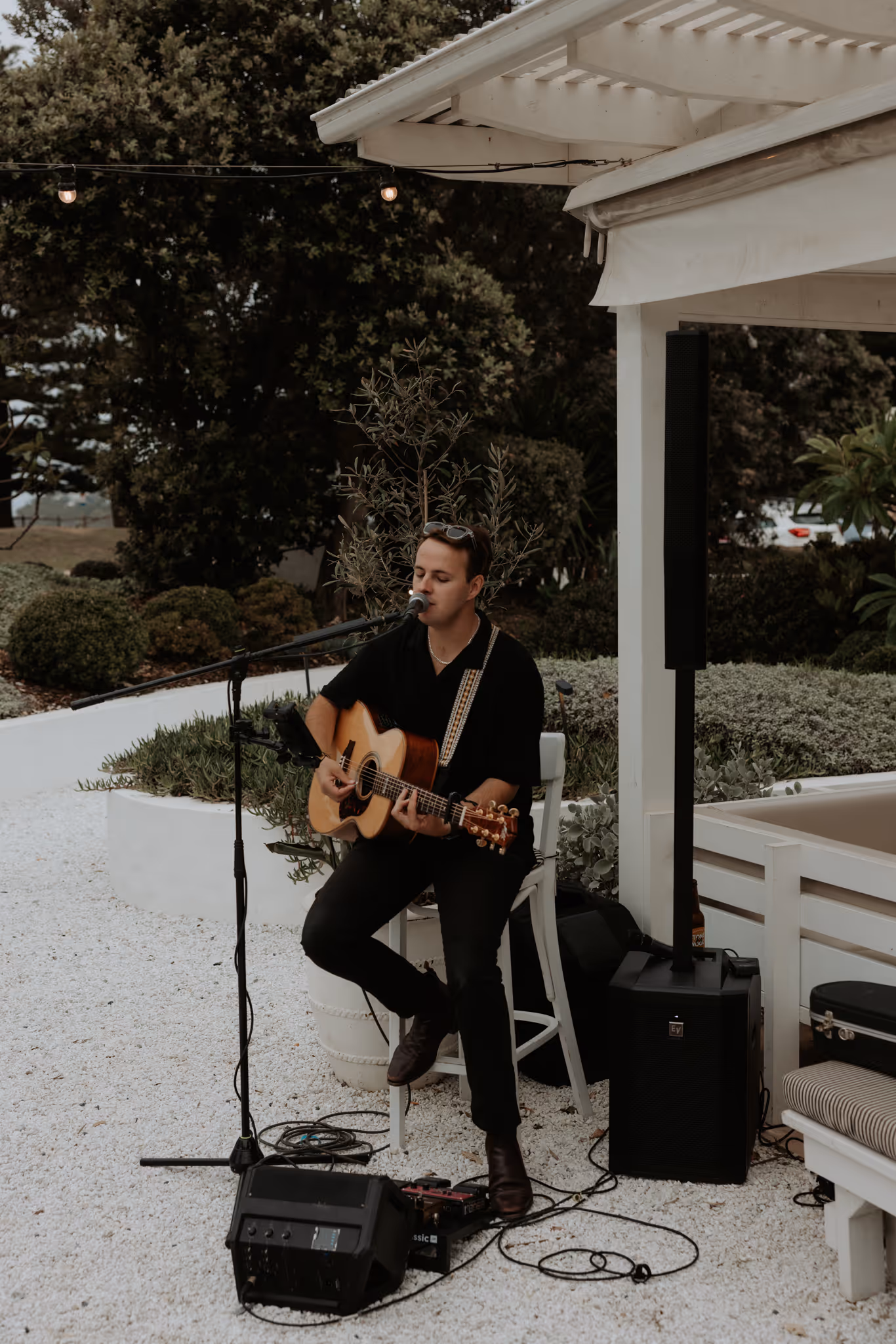Man sitting on a white chair playing acoustic guitar and singing into a microphone outdoors.