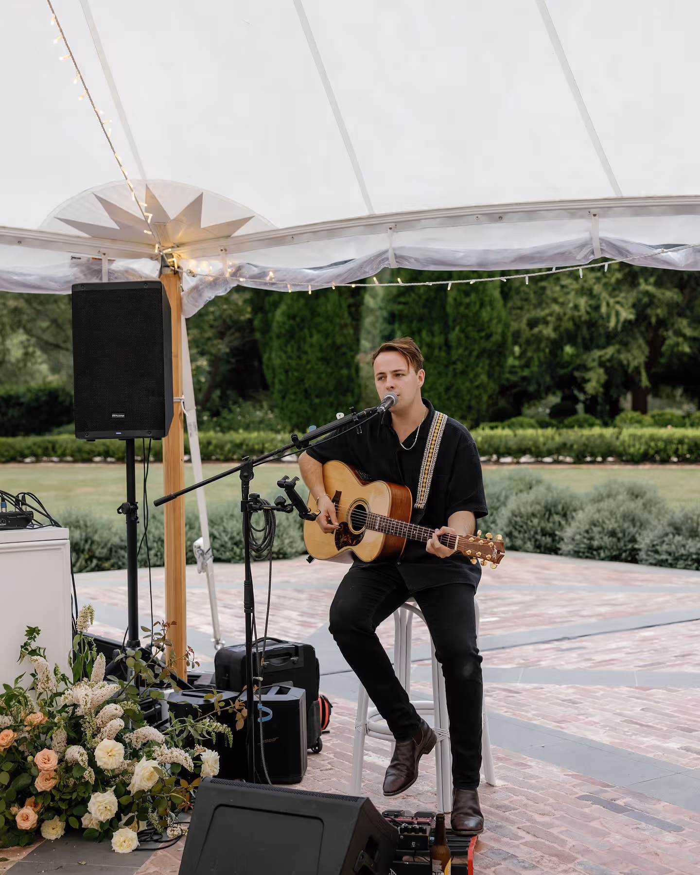 Man sitting on a white chair playing acoustic guitar and singing into a microphone outdoors.