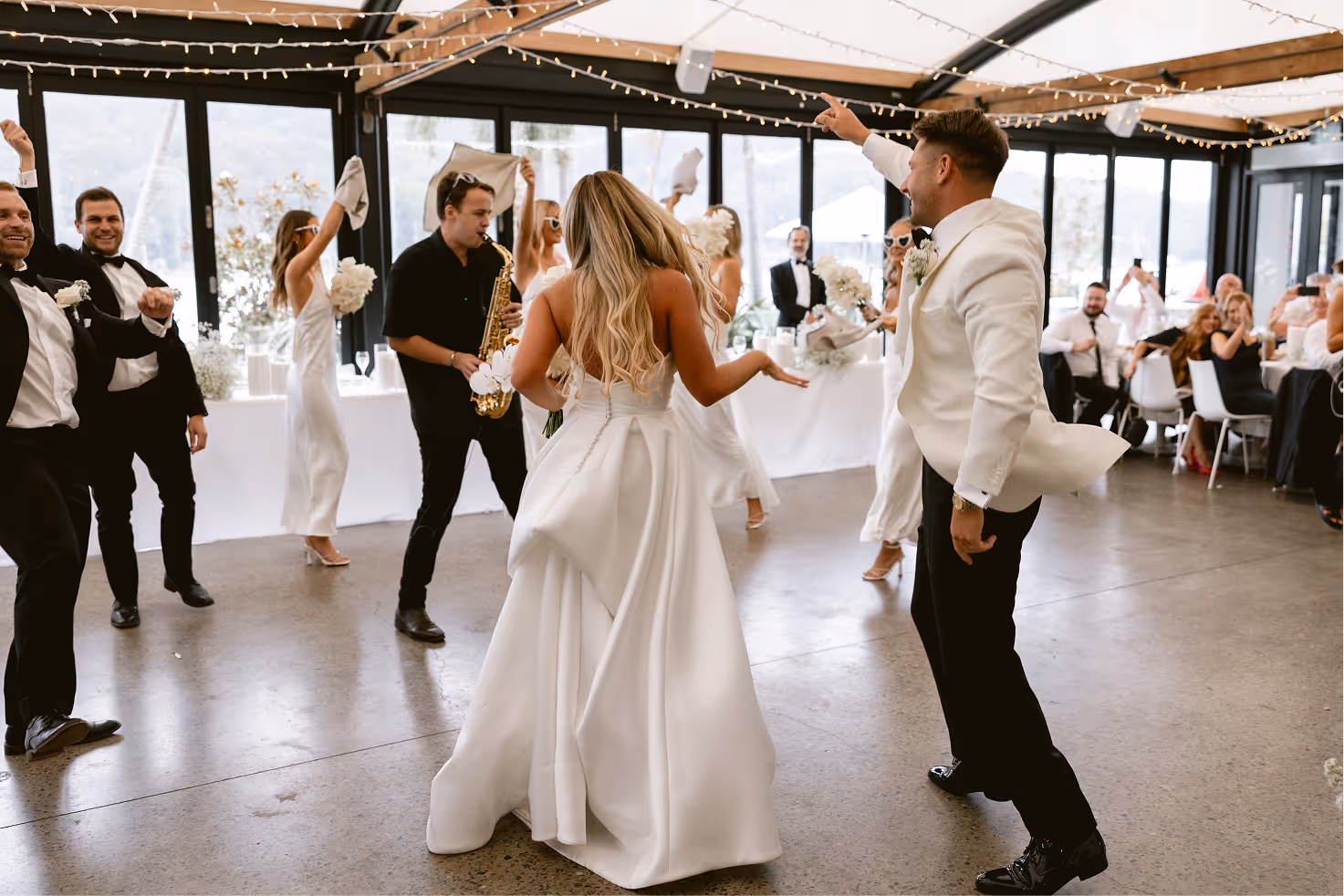 Bride and groom dancing indoors with a saxophone player and guests celebrating around them.