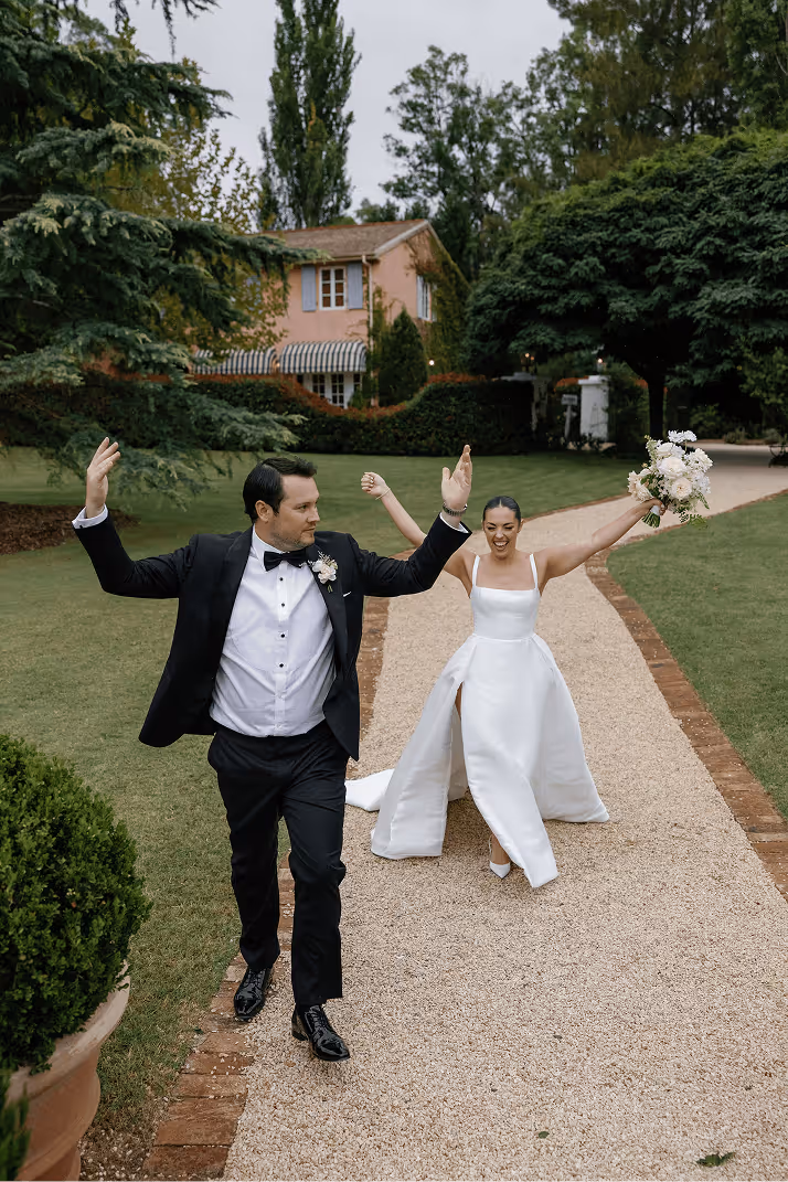 Bride and groom joyfully walking on a garden path with greenery and a pink house in the background.