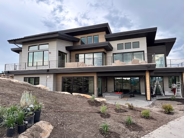 Modern two-story house with large windows, an upper balcony with seating, and a garden area under landscaping.
