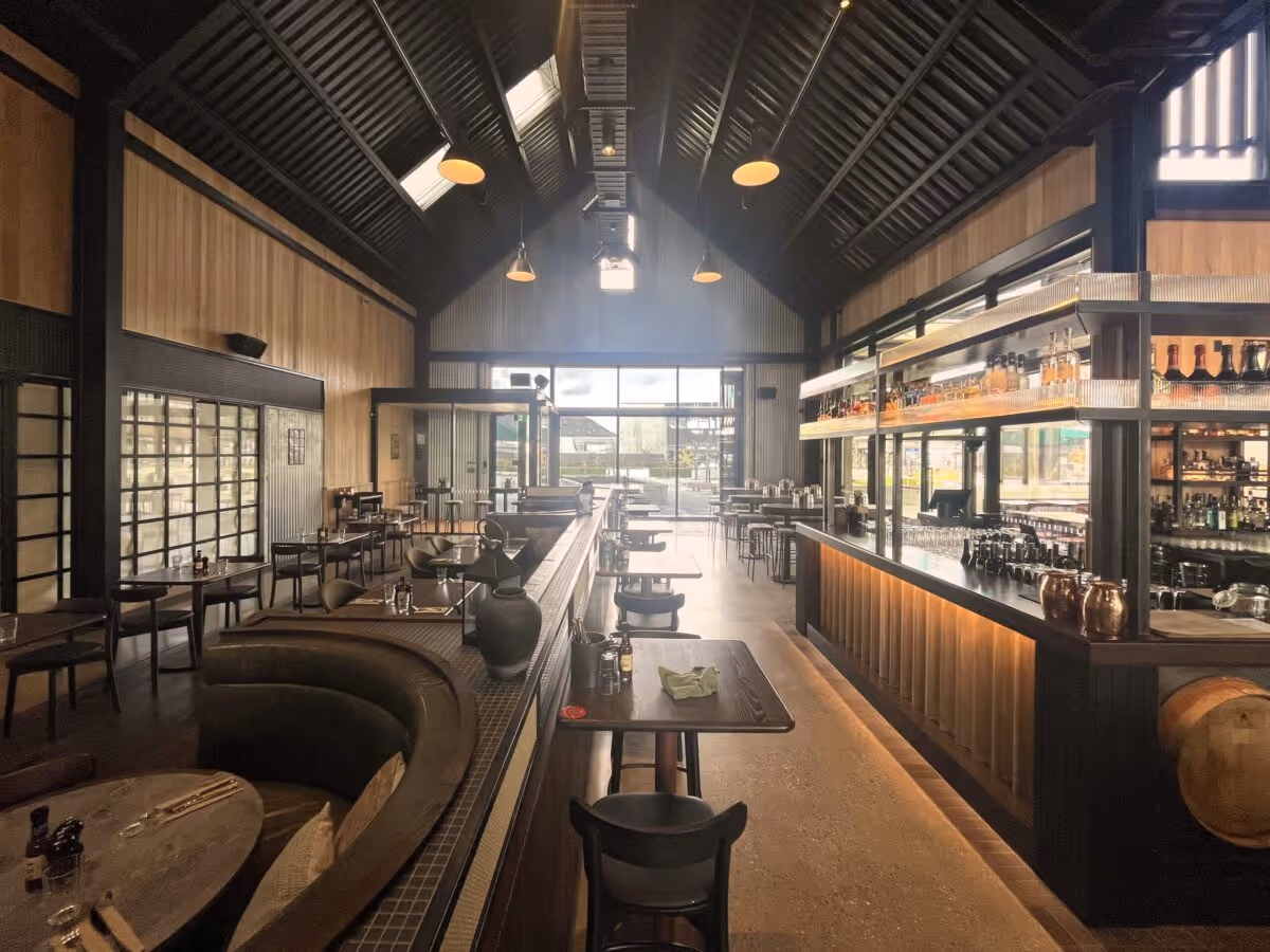 Modern restaurant interior with high vaulted ceiling, wooden panels, bar counter on right, and seating area with tables and chairs.