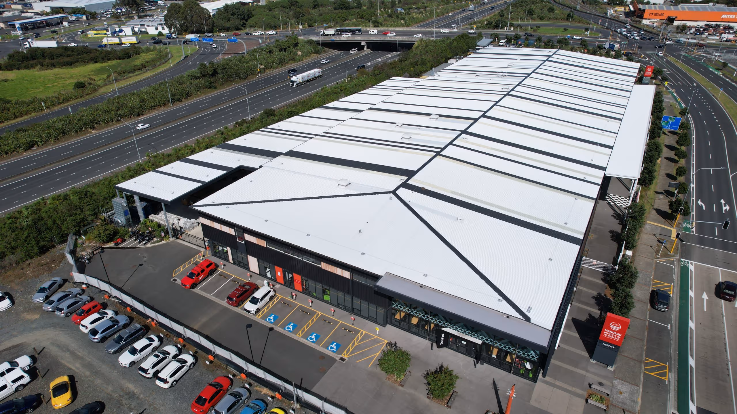 Aerial view of a large industrial warehouse with a white roof and parking spaces, located next to highways and surrounding roads.