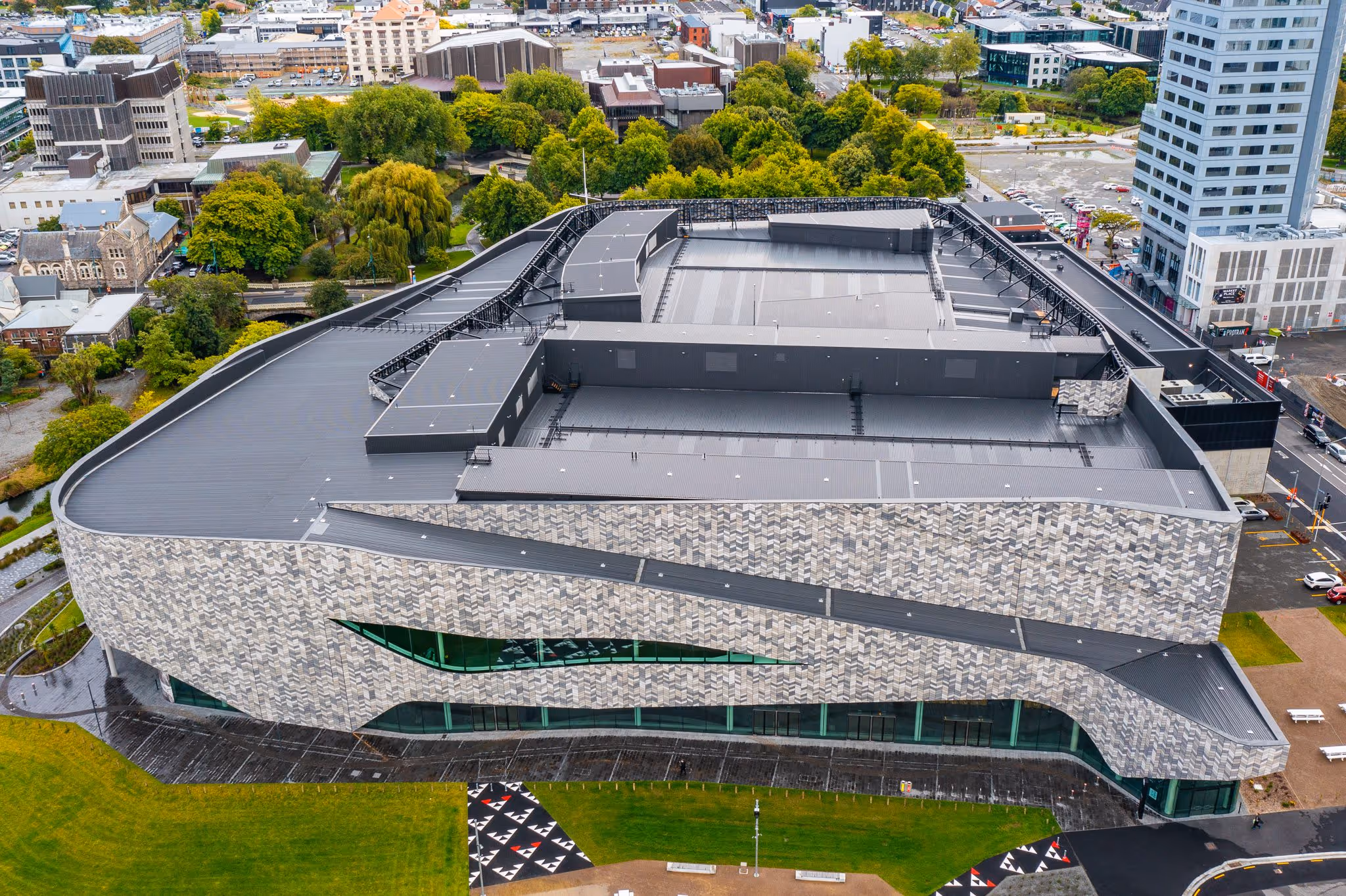 Aerial view of a modern arena with a patterned facade and curved glass windows surrounded by green trees and urban buildings.