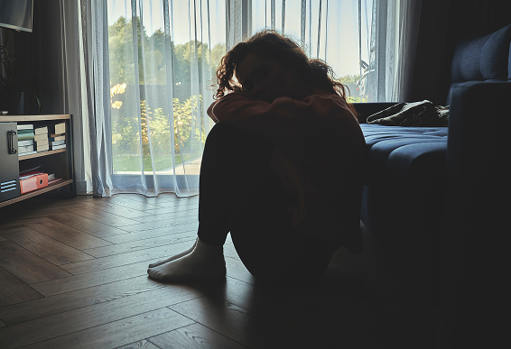 窓辺の床に座り、膝を抱えて沈んだ表情の女性。