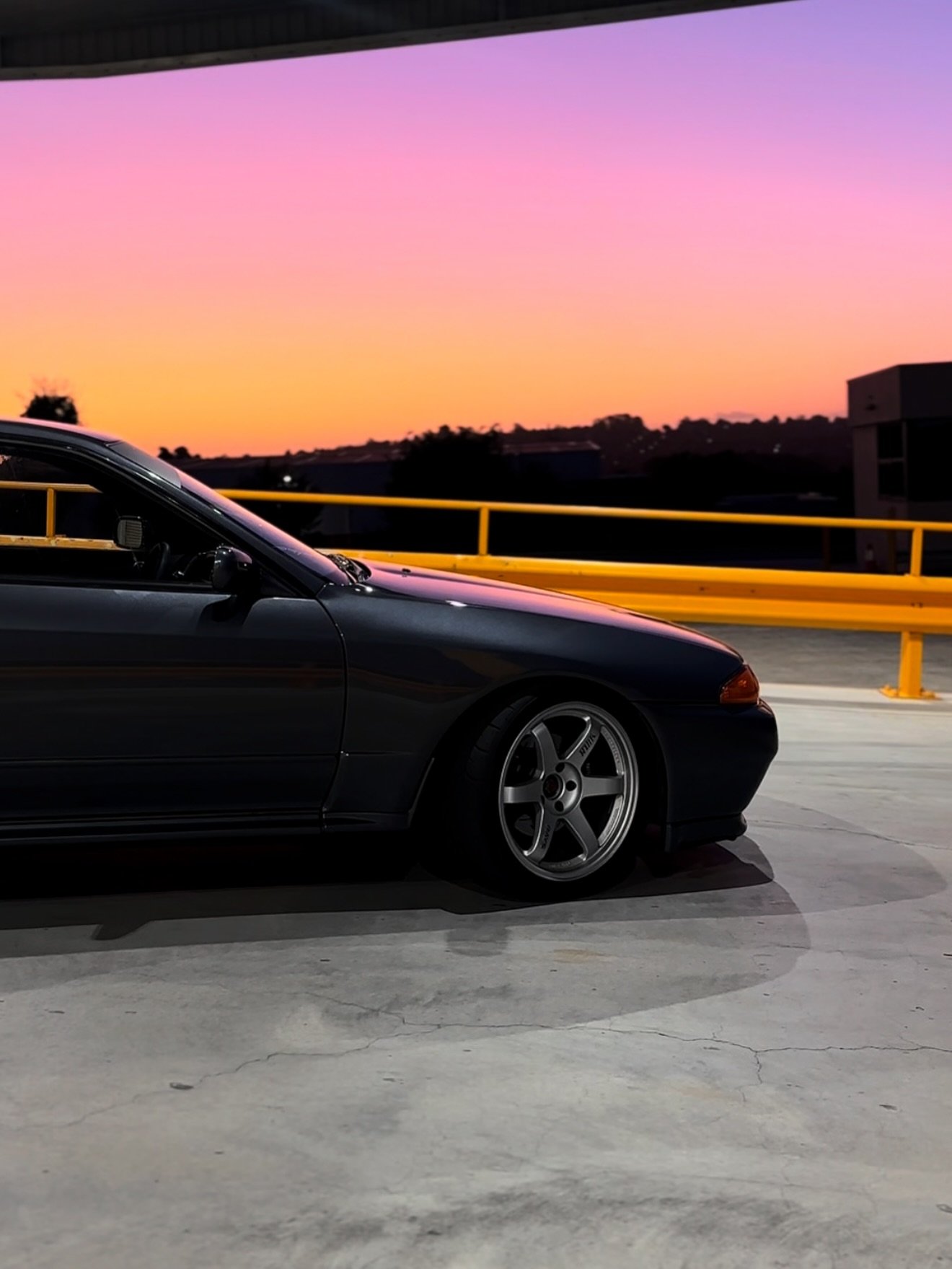 Side view of a dark car with lowered suspension and silver alloy wheels parked on concrete at sunset with a pink and orange sky.