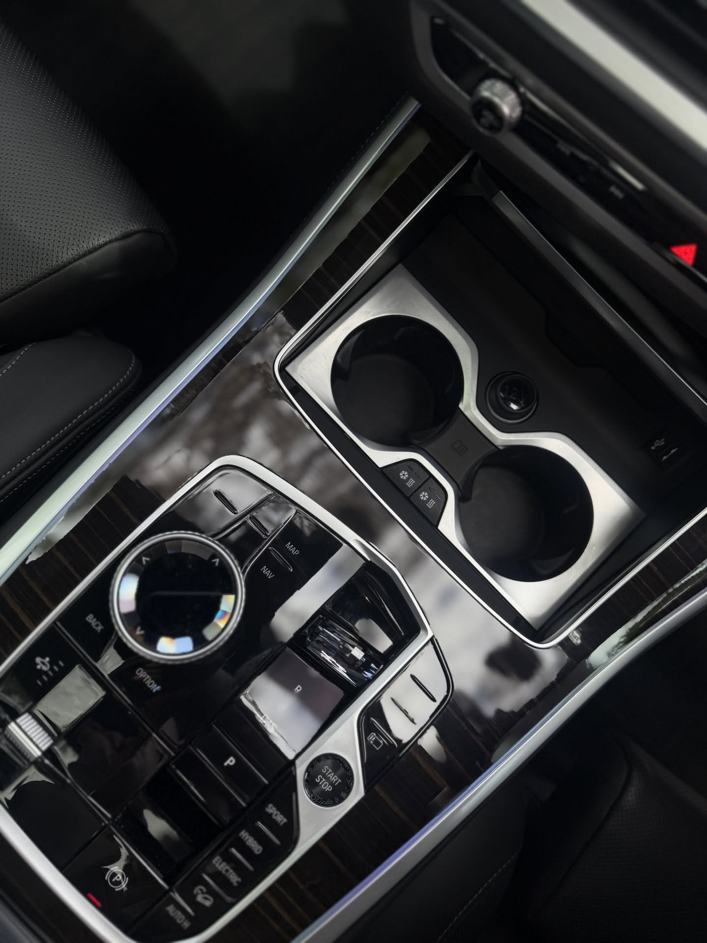 Close-up of a car's black and silver center console featuring a rotary control knob, buttons for navigation and driving modes, cup holders, and a start/stop engine button.