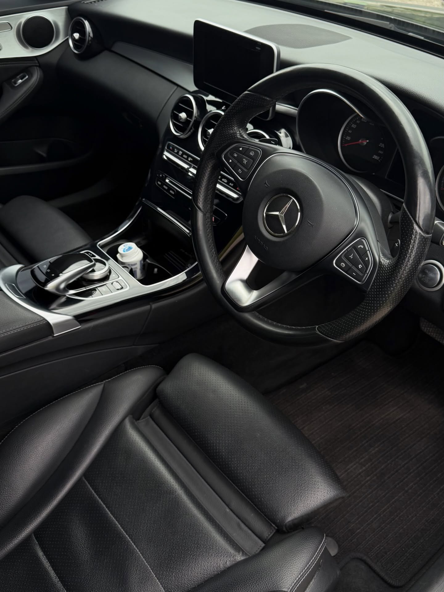 Driver's seat and dashboard interior of a black Mercedes-Benz car, showing steering wheel, center console, and infotainment screen.