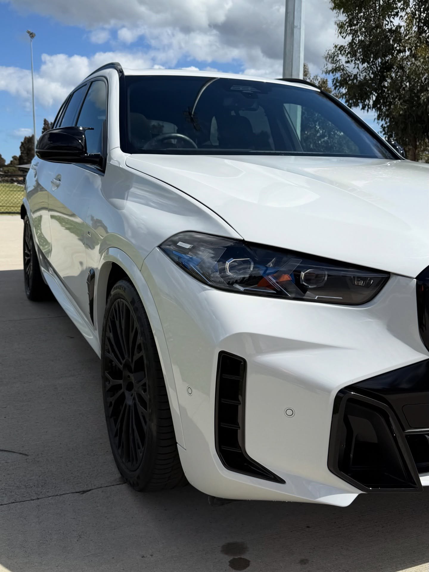 Front side view of a white luxury SUV with black rims and tinted windows parked on a concrete surface under a partly cloudy sky.