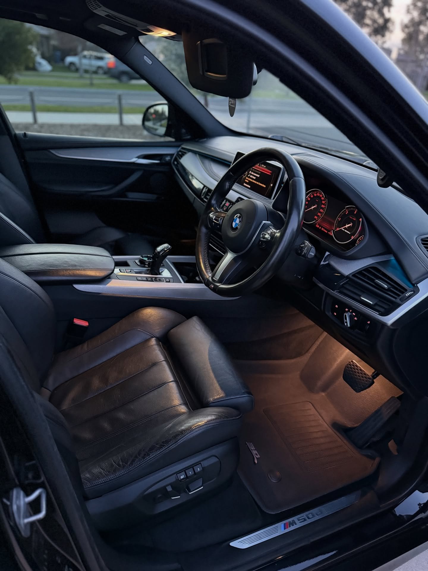 Interior of a black BMW M500i showing the driver's seat, steering wheel, dashboard, and illuminated footwell.