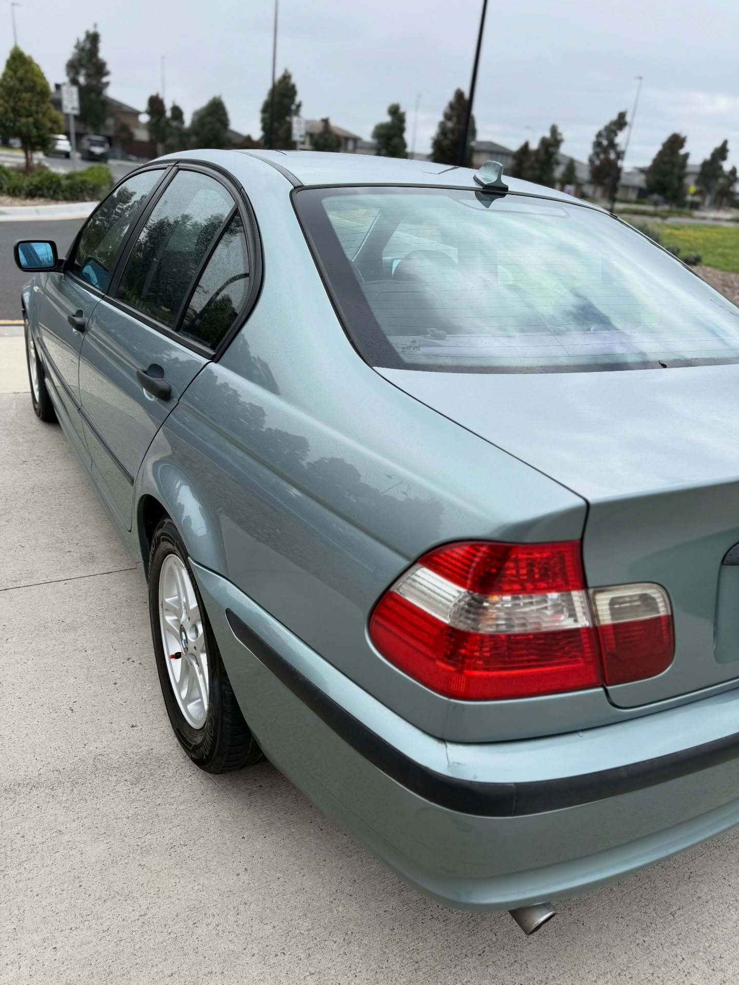 Light blue sedan parked on a concrete driveway, viewed from the rear left side showing the tail light and rear window.
