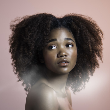 Portrait of a young woman with natural curly hair looking to the side against a soft pink background.