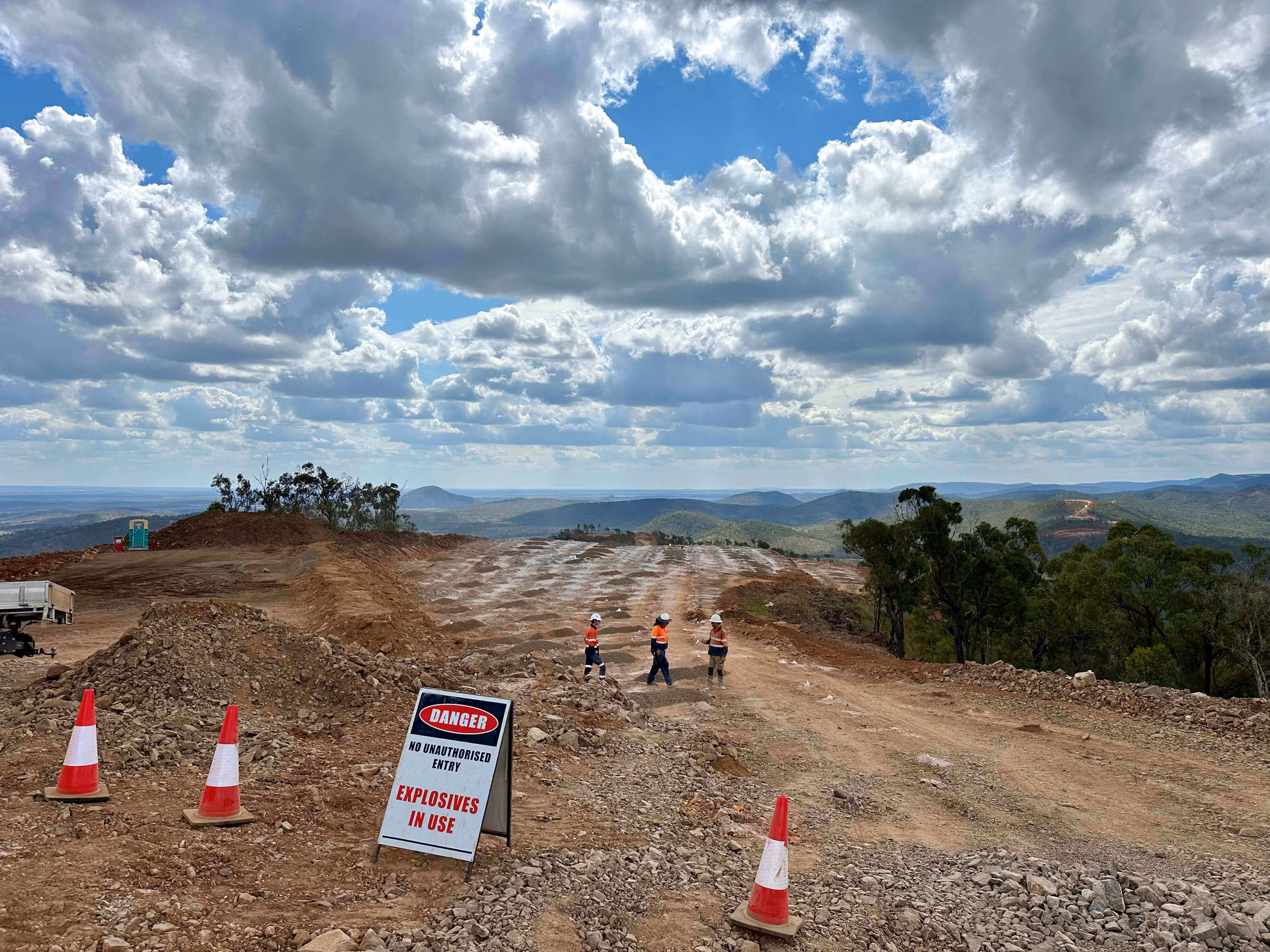Construction site on a hill with three workers in high-visibility gear near a sign reading 'Danger No Unauthorised Entry Explosives In Use', under a partly cloudy sky.
