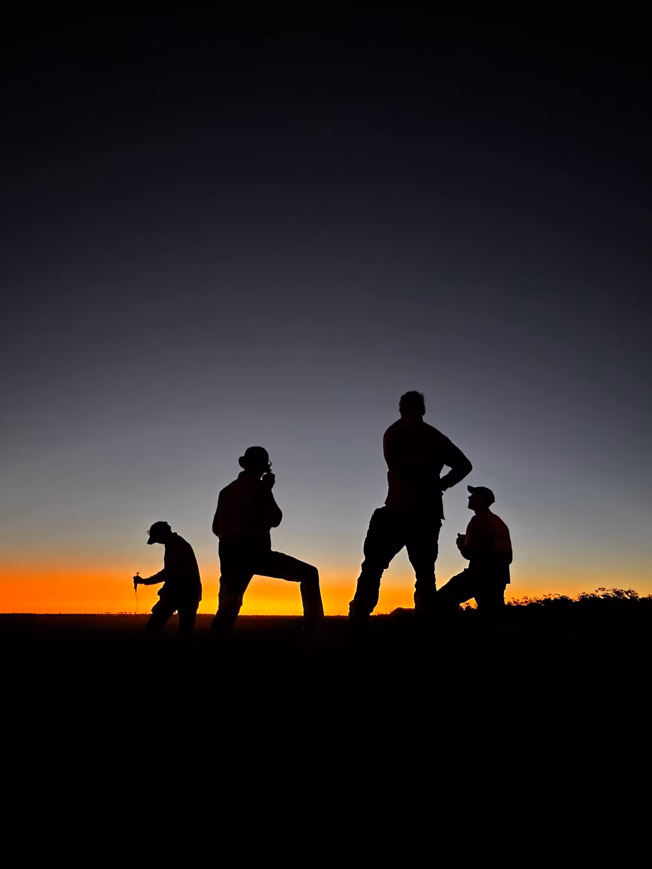 Silhouettes of four people standing and sitting against a vibrant orange and darkening blue sunset sky.