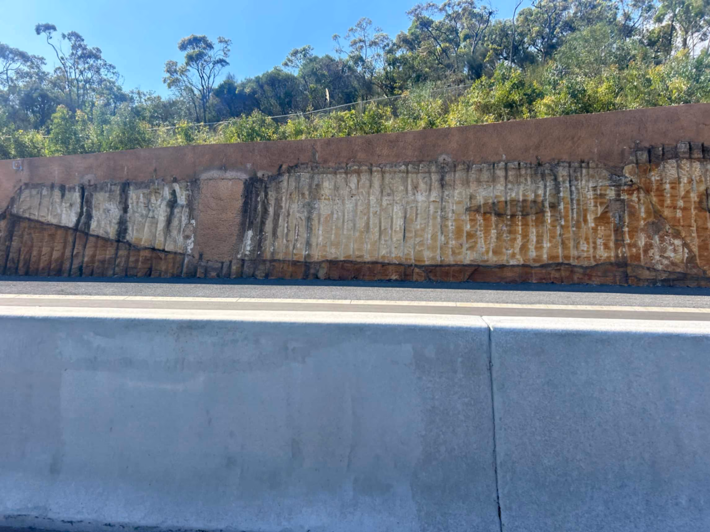 Exposed rock layers and vertical drill lines in a roadside cut with trees above the embankment.