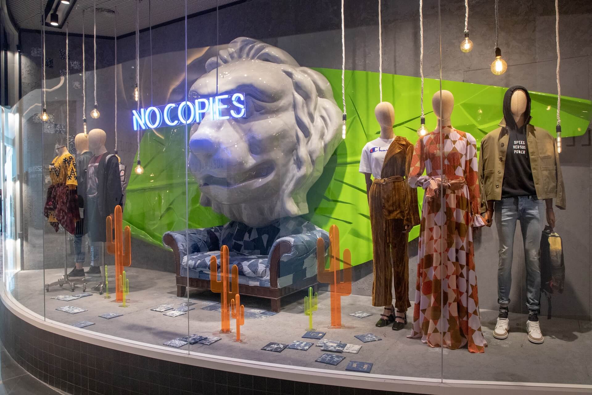 Fashion store window display with mannequins, lion statue sculpture, and neon "NOCOPIES" sign.