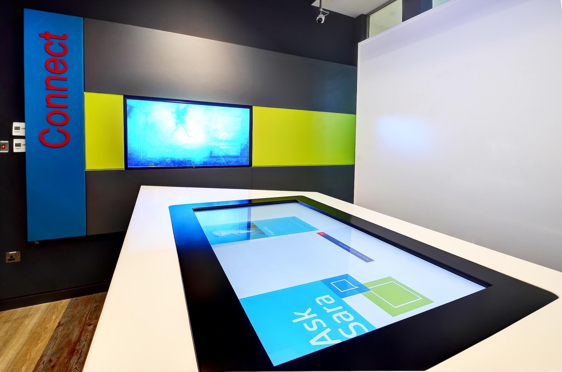 Modern exhibit space with interactive touch display, wall screens, and blue Comtech signage