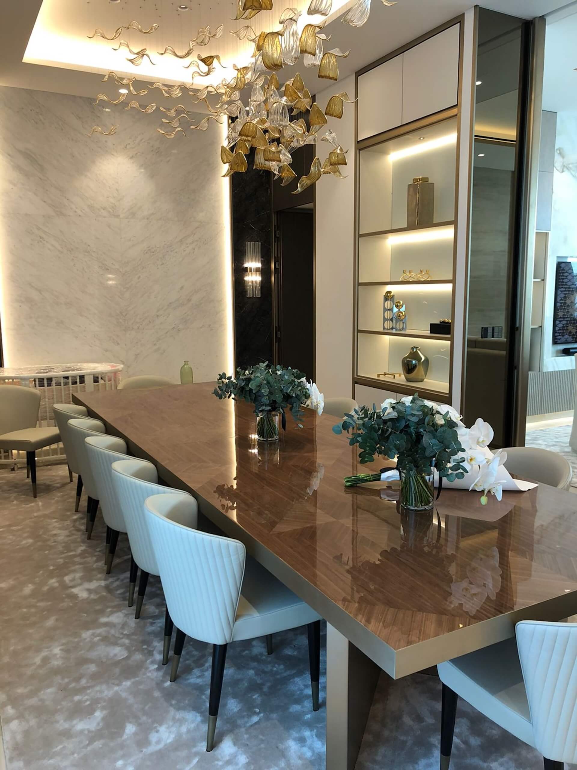 Modern dining room with long wood table, white chairs, decorative chandelier, and illuminated shelving.