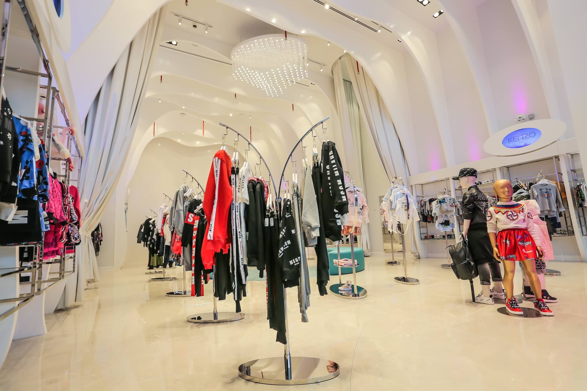 Modern fashion retail store interior with clothing displays, mannequins, and elegant white ceiling with chandelier.