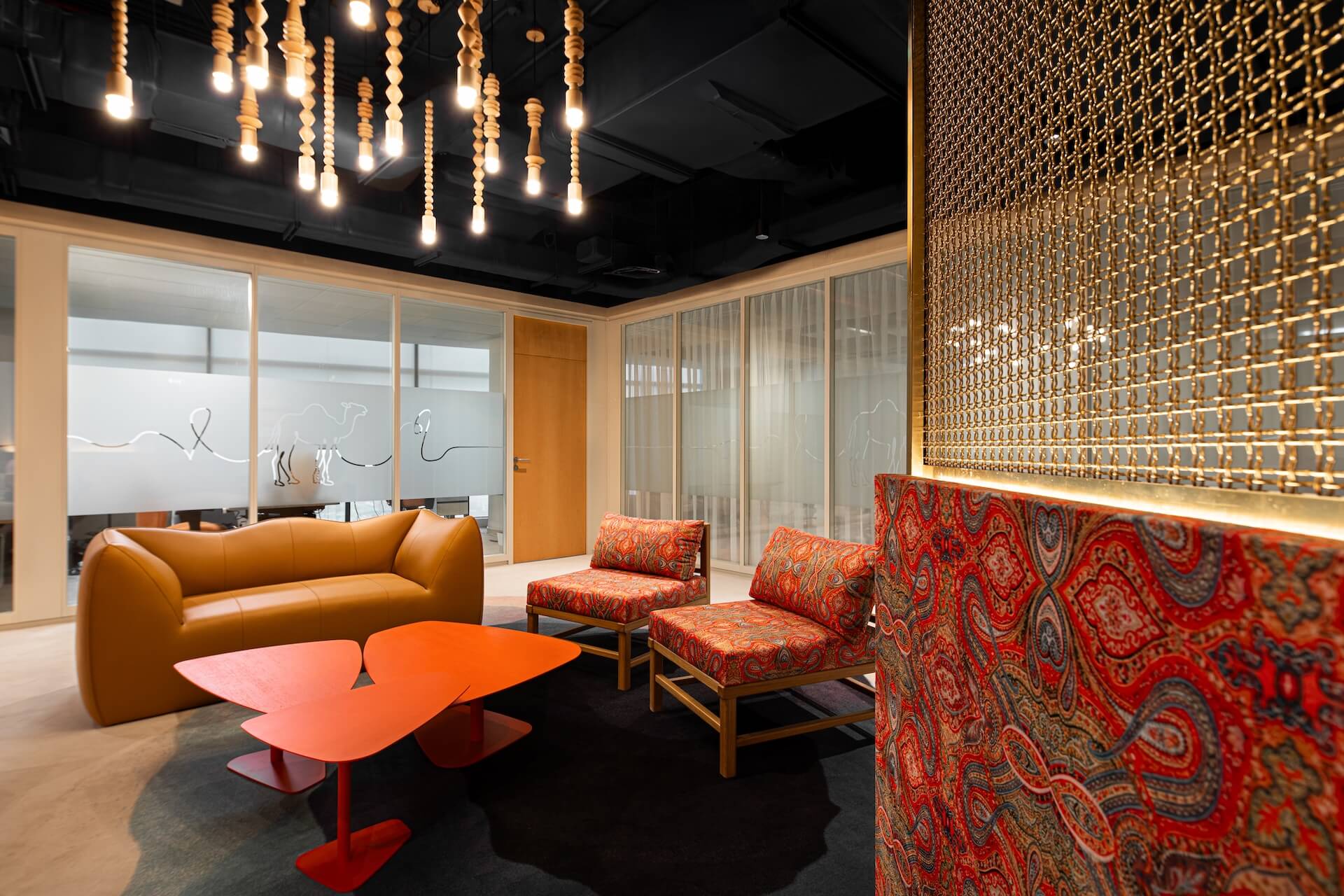 Modern office lounge with leather sofa, patterned chairs, red table, and gold geometric wall partition.