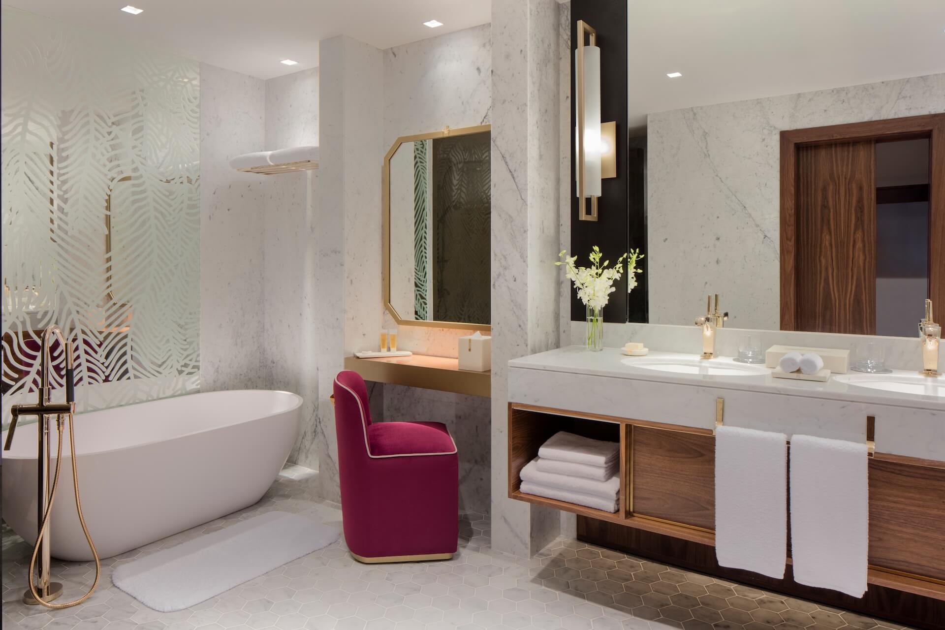 Modern luxury bathroom with marble walls, freestanding tub, dual vanity, and burgundy accent chair.