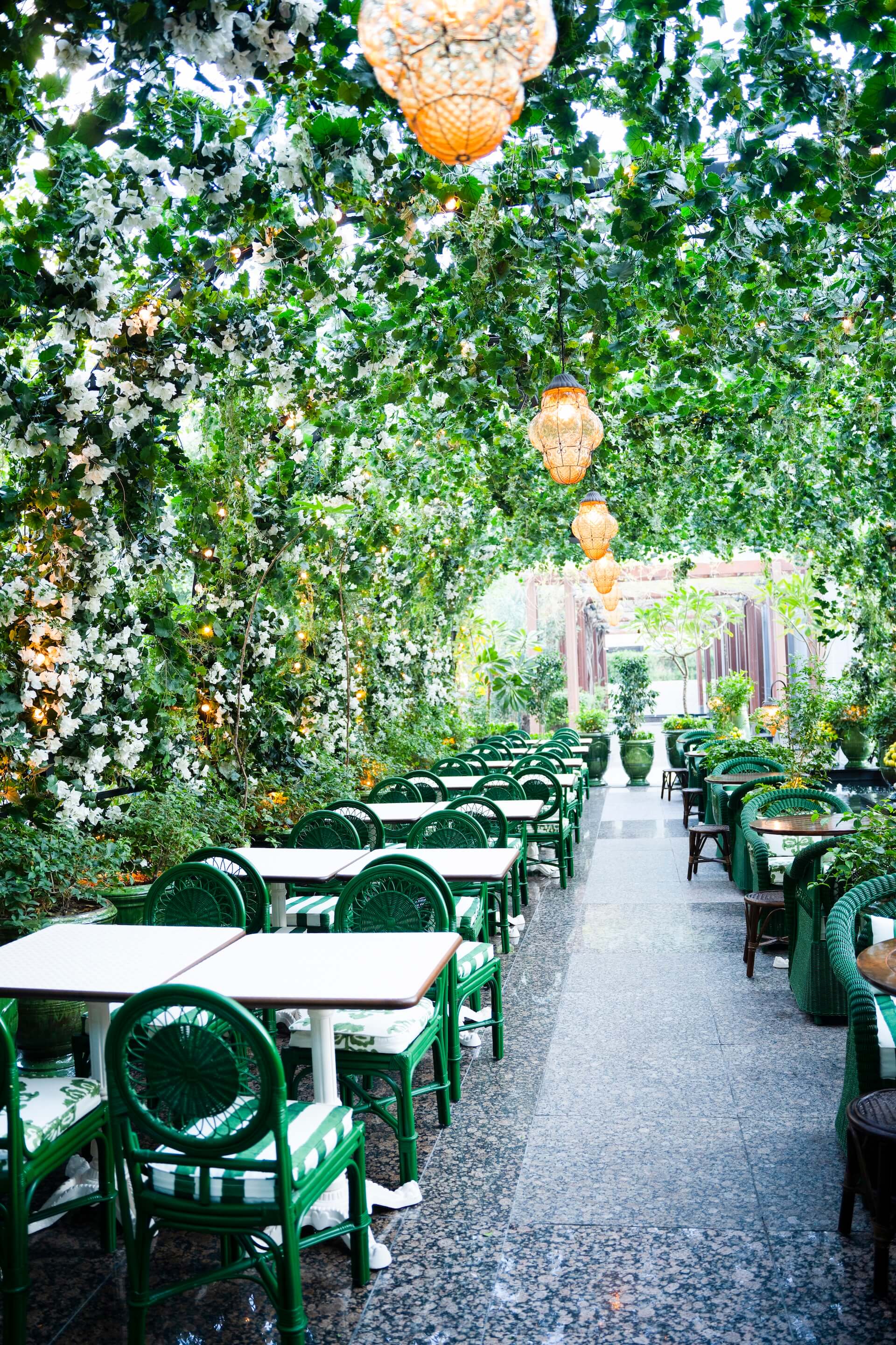 Lush outdoor restaurant patio with green chairs, white tables, cascading ivy and hanging lanterns overhead