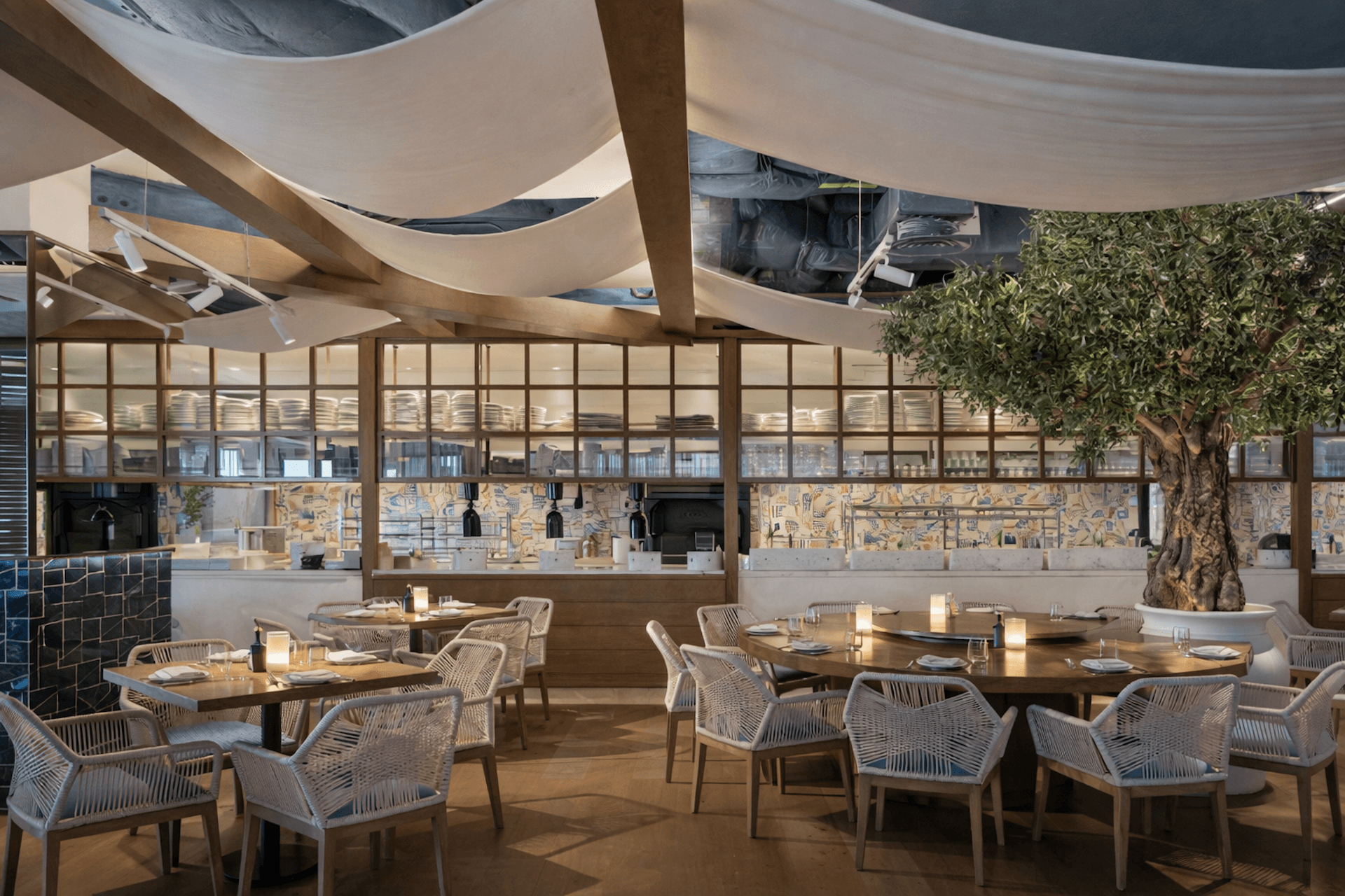 Modern restaurant interior with cream fabric canopy, wooden tables, woven chairs, and an olive tree.