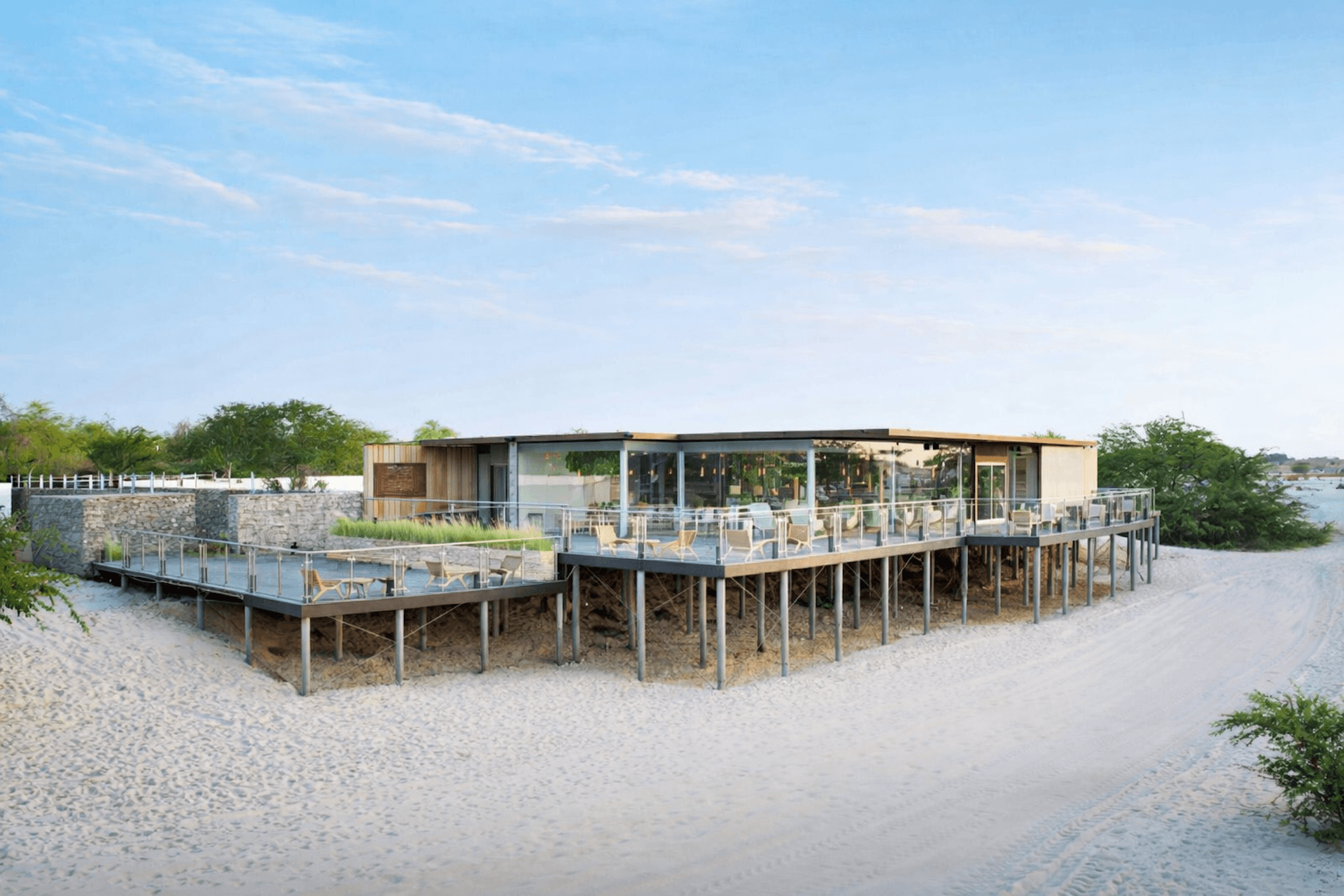 Modern beach house on stilts with deck overlooking sandy shore and water.