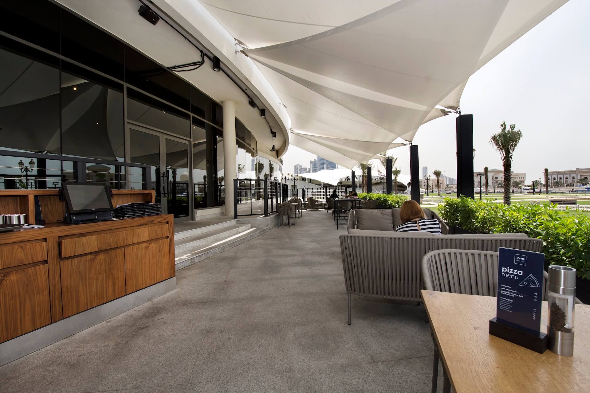 Modern outdoor restaurant patio with white sail shade, seating, bar counter, and palm trees overlooking plaza