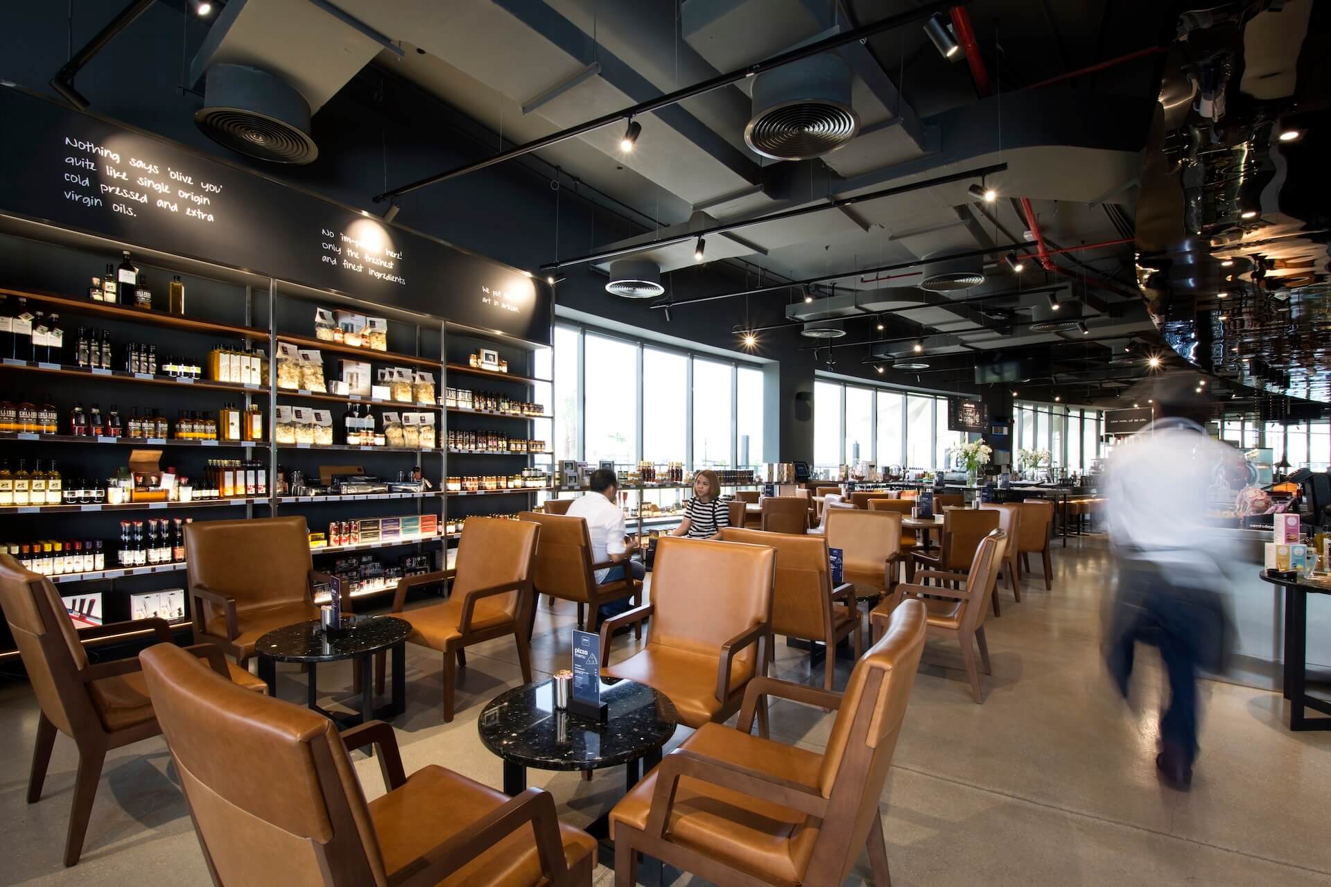 Modern boutique shop interior with tan leather seating, shelved products, and natural lighting from large windows.