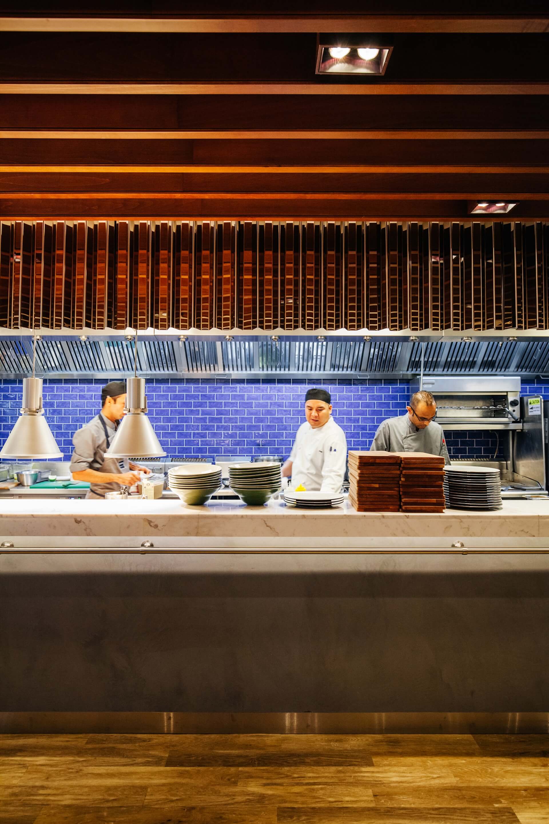 Four chefs work in modern open kitchen with blue tile backsplash and stacked white plates