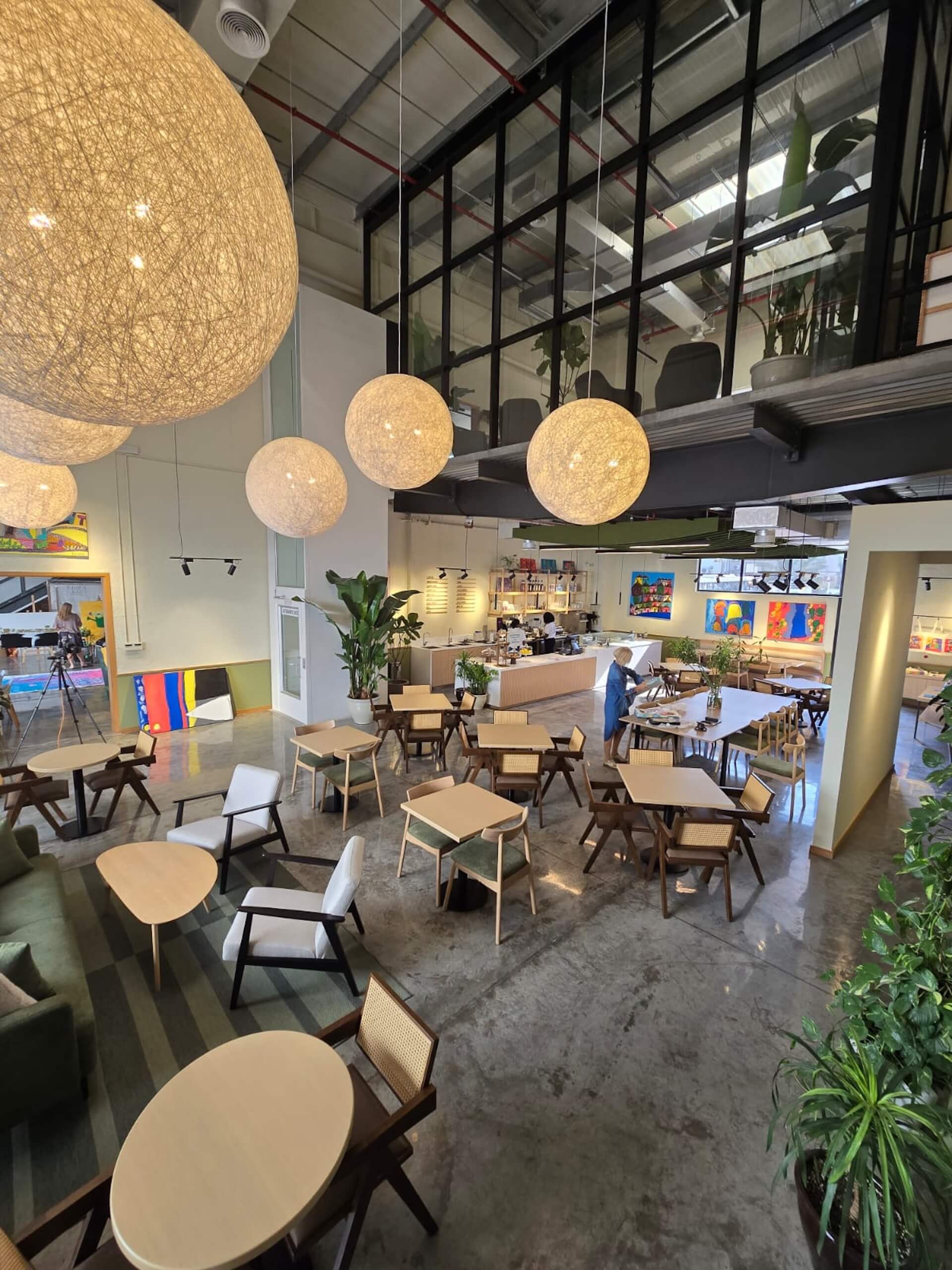 Modern café interior with pendant sphere lights, wooden tables, plants, and glass-enclosed office space